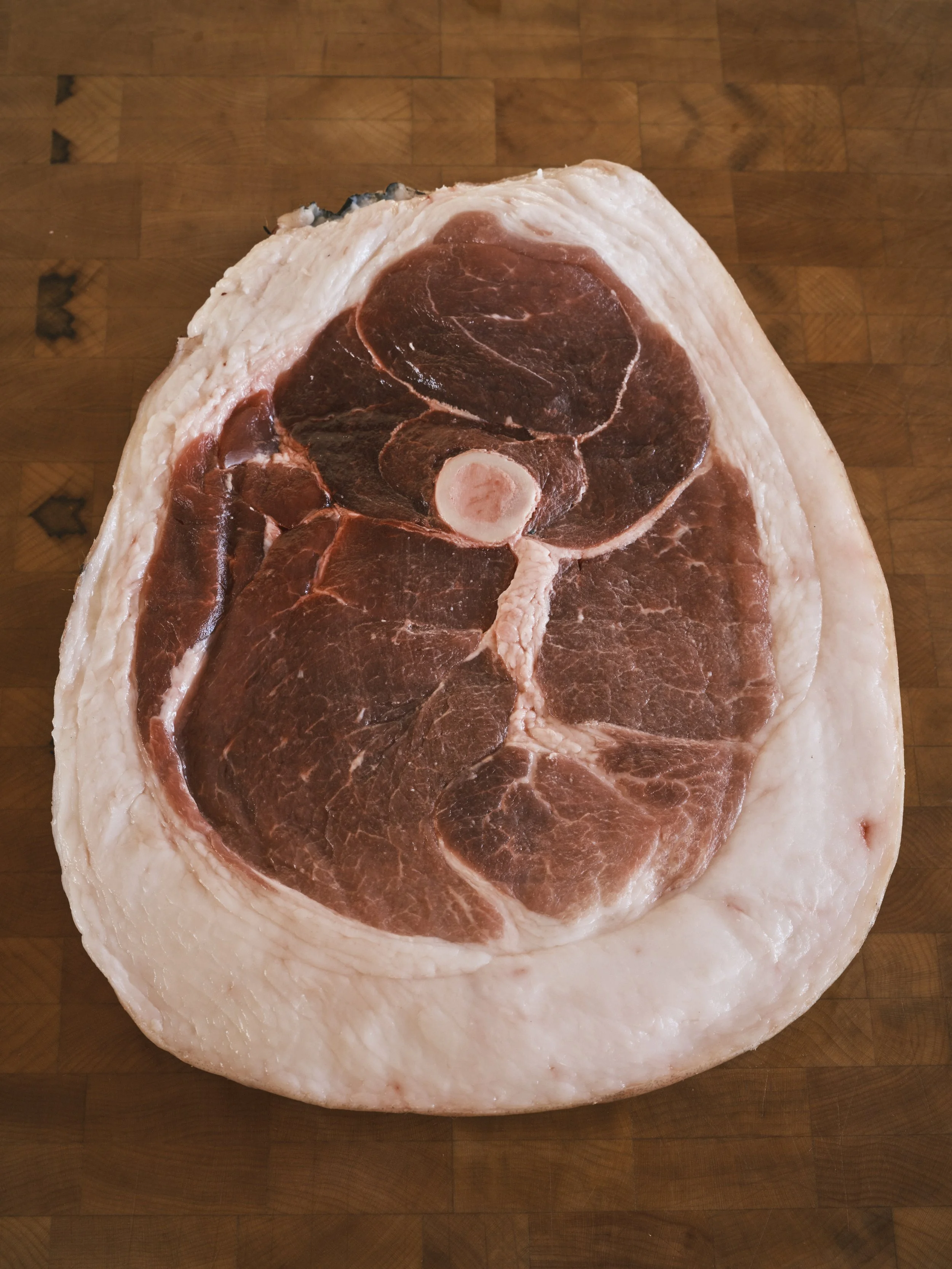 A large cut of raw ham, showing the bone and marbled meat, placed on a wooden cutting board.
