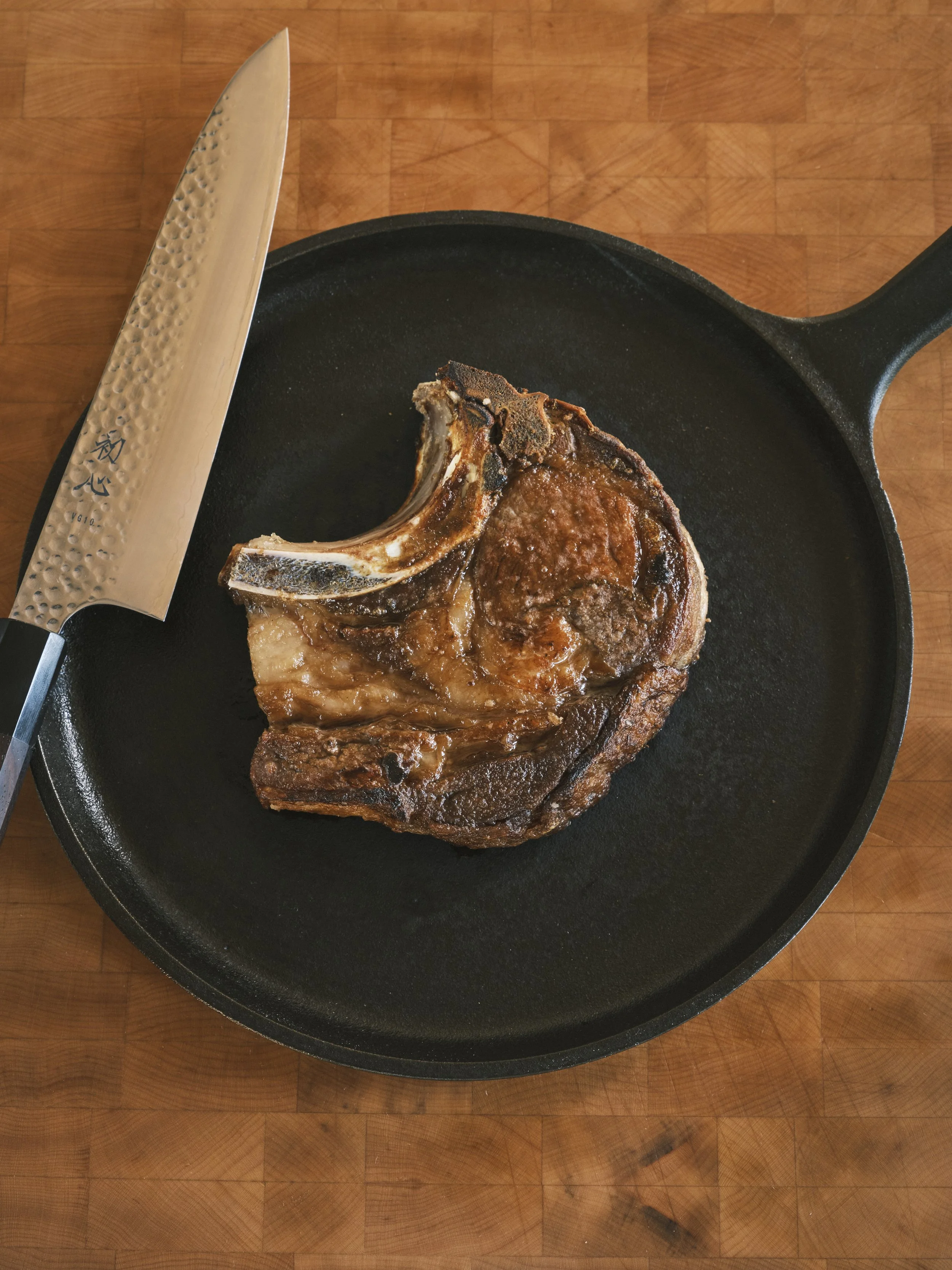A cooked beef short rib in a black cast iron skillet with a Japanese-style chef's knife nearby, on a wooden surface.