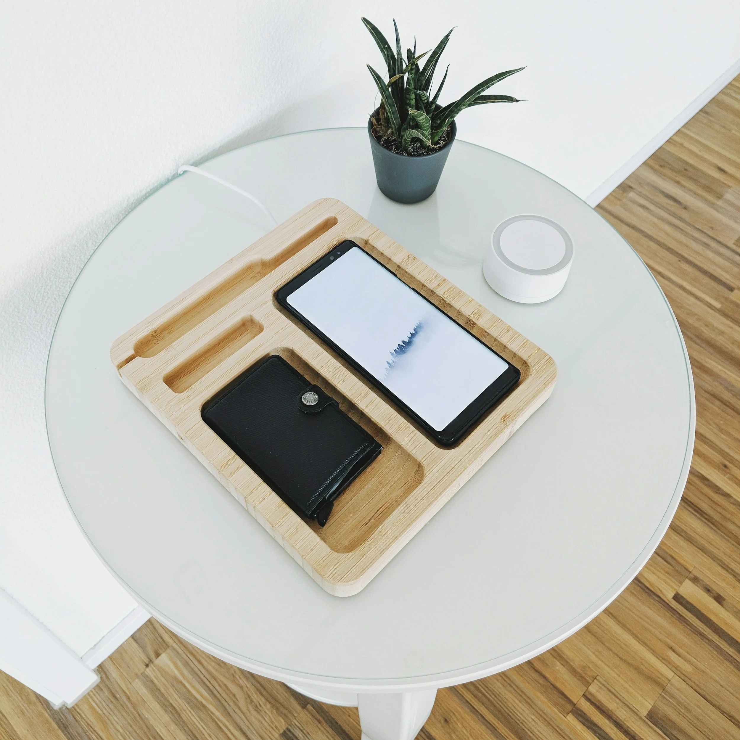 Round white table with a wooden tray holding a smartphone and a wallet, a potted plant, and a white smart speaker on a hardwood floor.