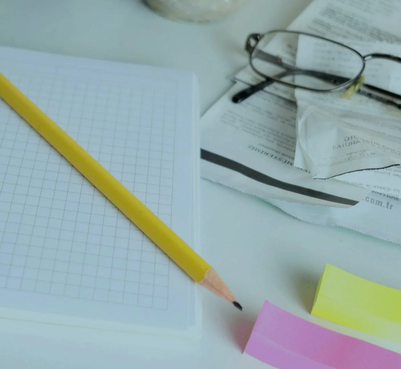A desk with a stationery set including yellow pencil, sticky notes in pink and yellow, a gridded notepad, a pair of glasses, and various papers and documents.