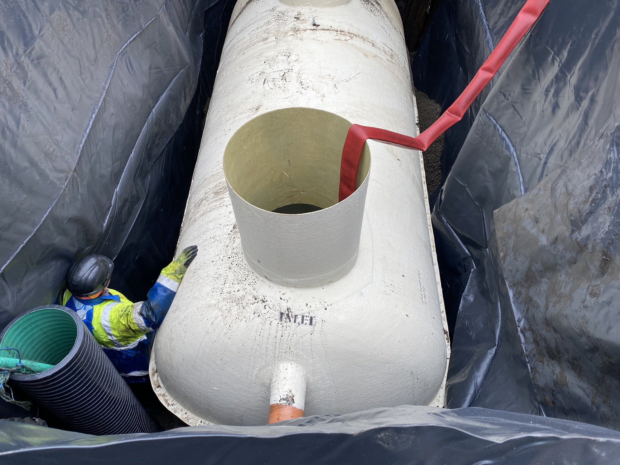 A construction worker wearing a hard hat and safety gear is working on a large cylindrical pipe, with black pipes or hoses stacked nearby, and black plastic sheeting surrounding the area.