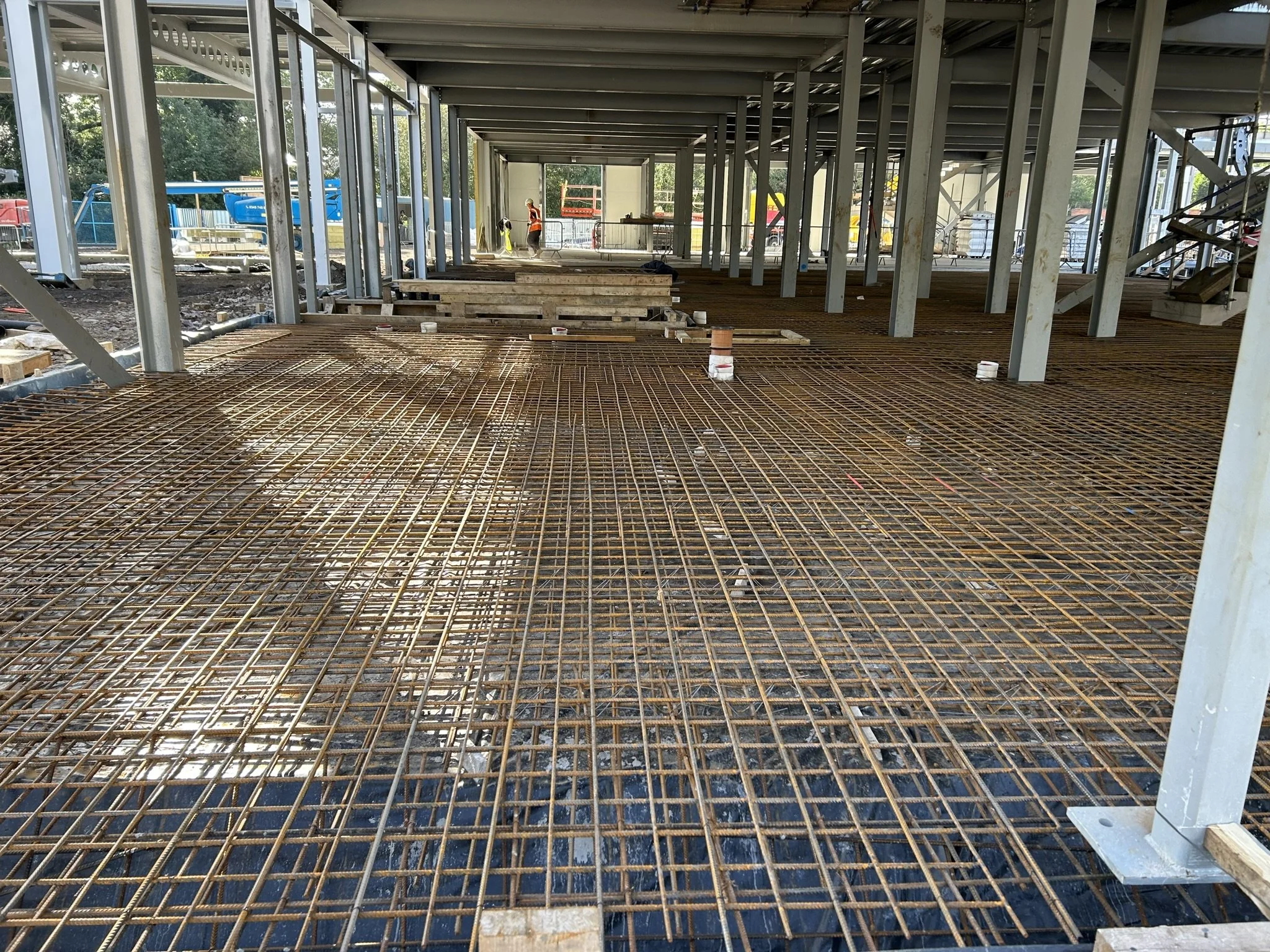 Construction site showing a steel rebar grid for the floor of a building, with workers in the background.