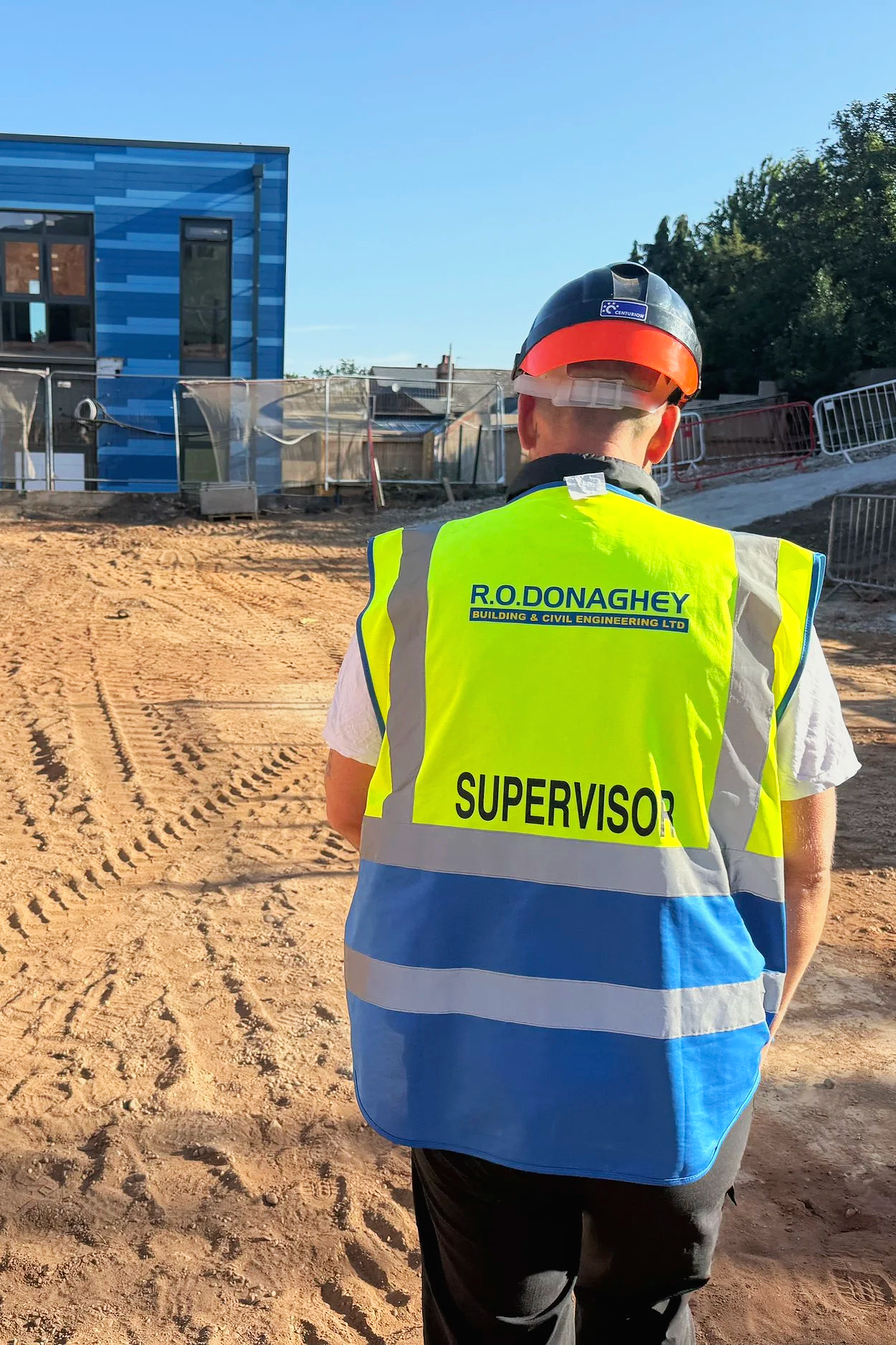 A construction supervisor wearing a high-visibility vest with the name R.O. Donaghey and the word 'Supervisor' on the back, standing on a construction site with a partially built modern blue building in the background.