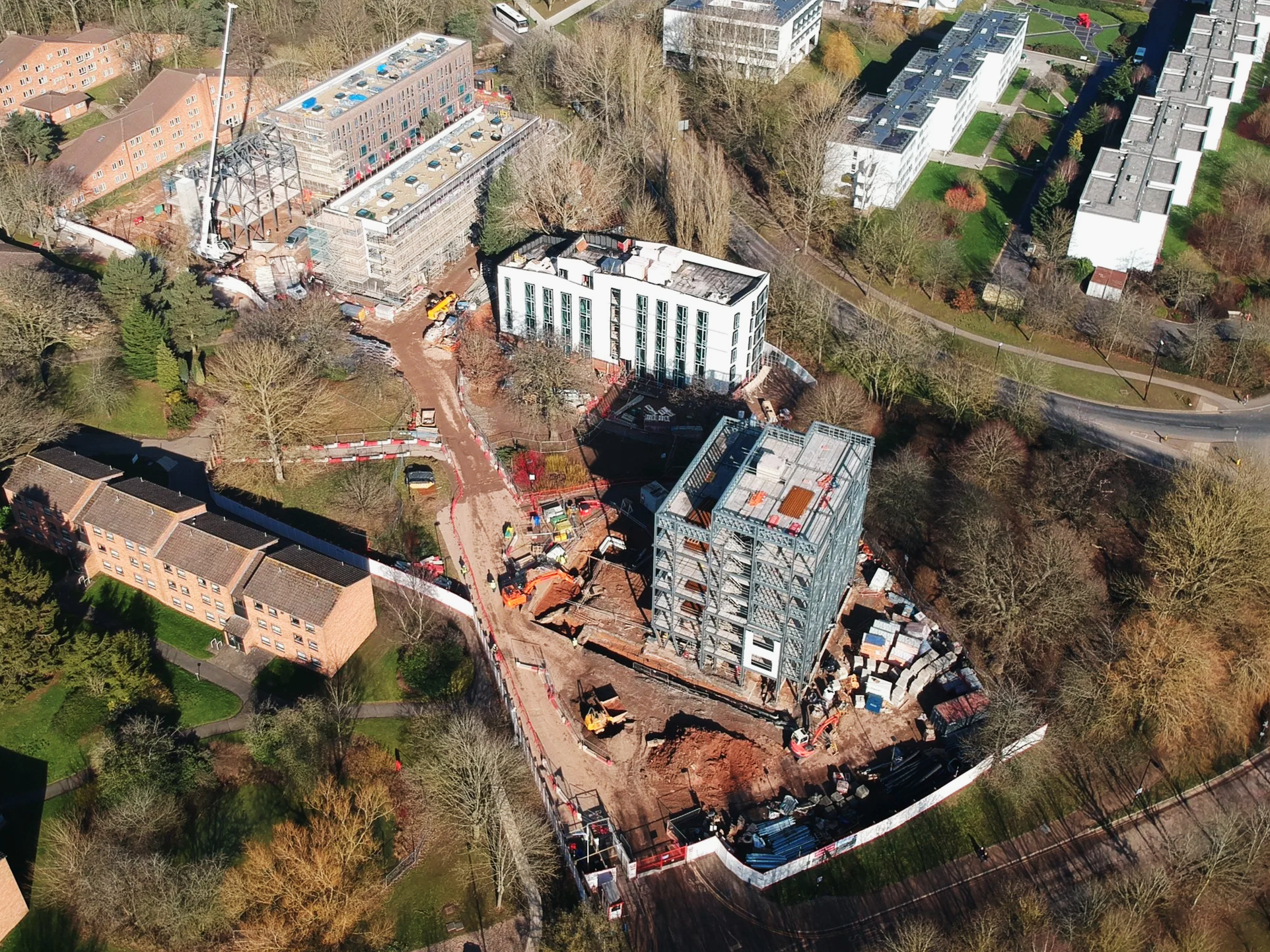 Bird's eye view of a construction site with two buildings under construction, surrounded by trees and residential buildings, with construction vehicles and materials on site.