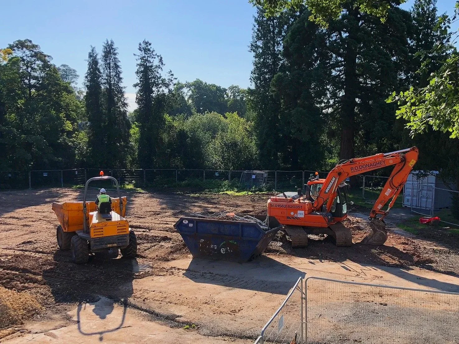 Construction site with a worker on a roller machine and an orange excavator working on the ground, surrounded by trees.