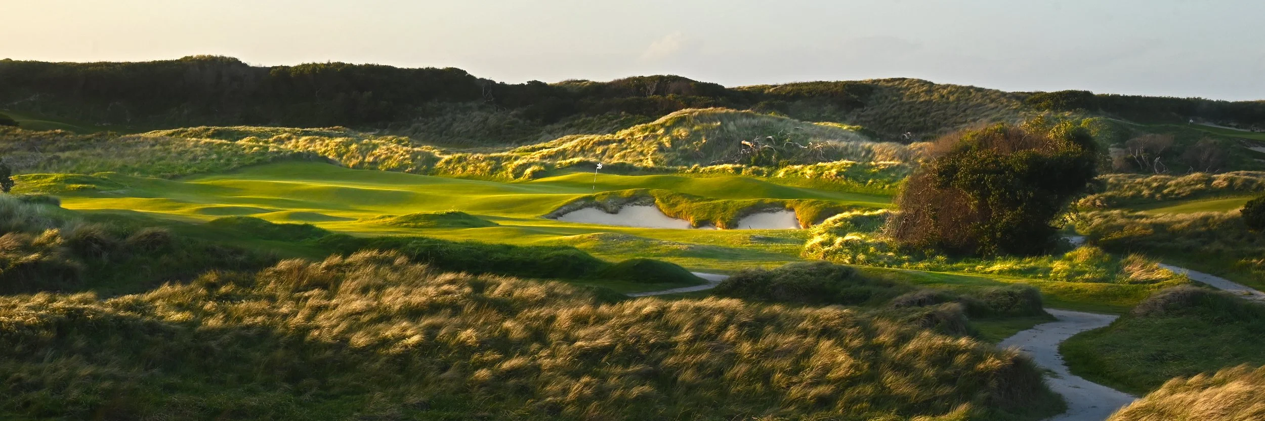 A scenic golf course with greens, sand bunkers, and a flagstick, surrounded by rolling hills and grassland.