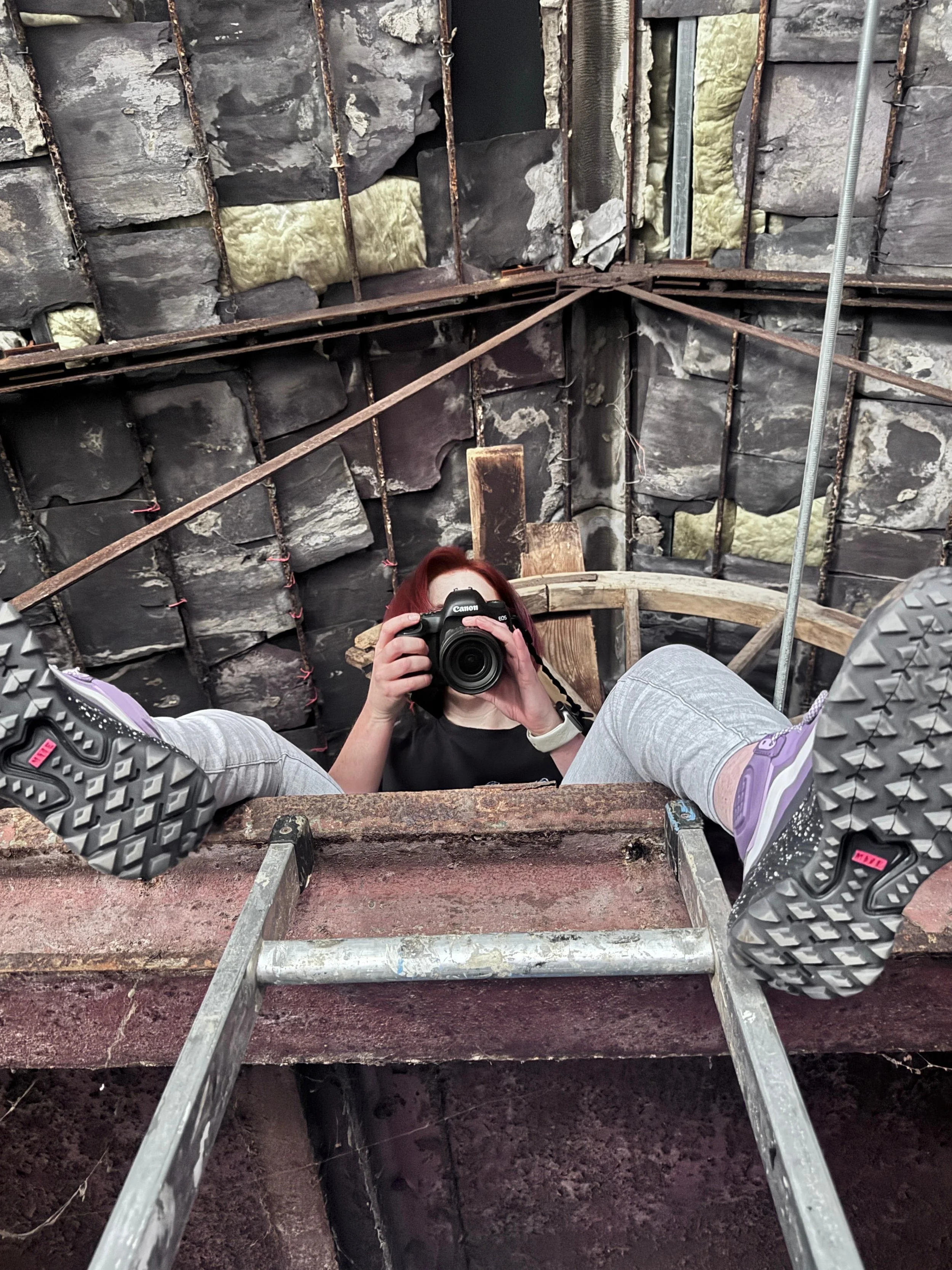 A person taking a photo with a camera while lying on a ladder in an unfinished or under-construction room with stone walls.