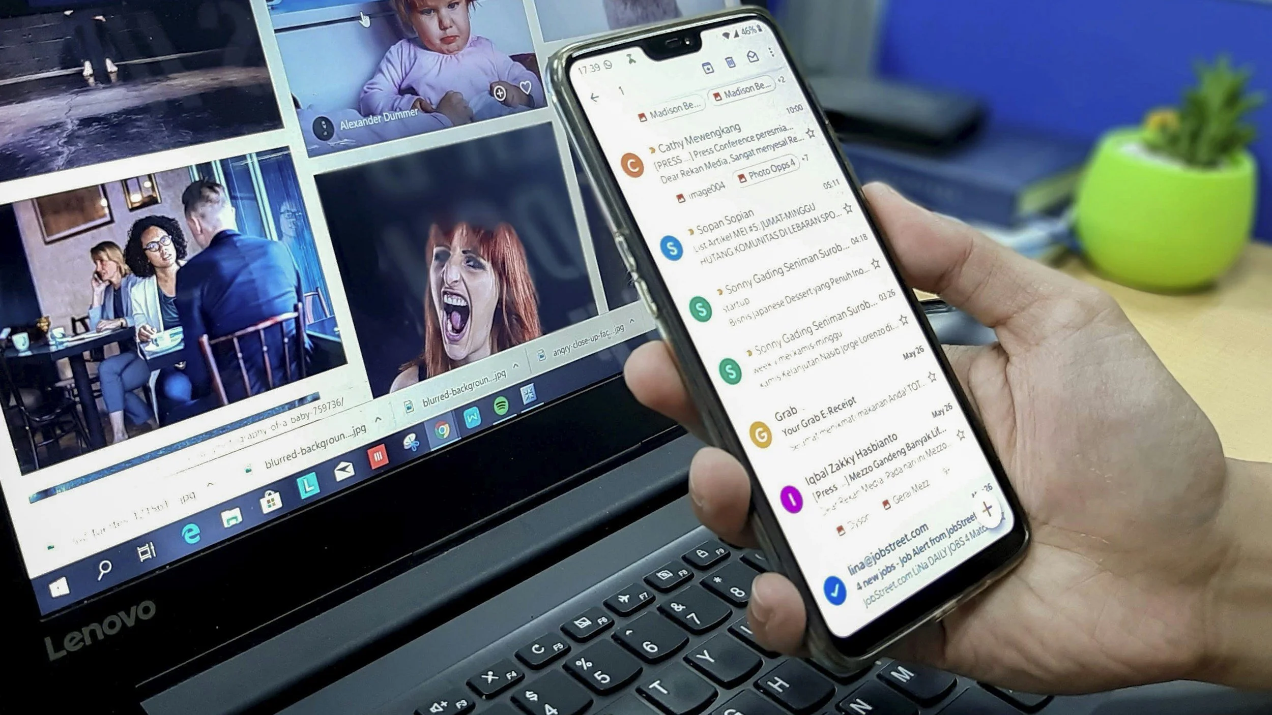 A person holds a smartphone displaying an email inbox, with a laptop in the background showing a grid of images including a child, a woman shouting, and a man talking to two women during a meal.