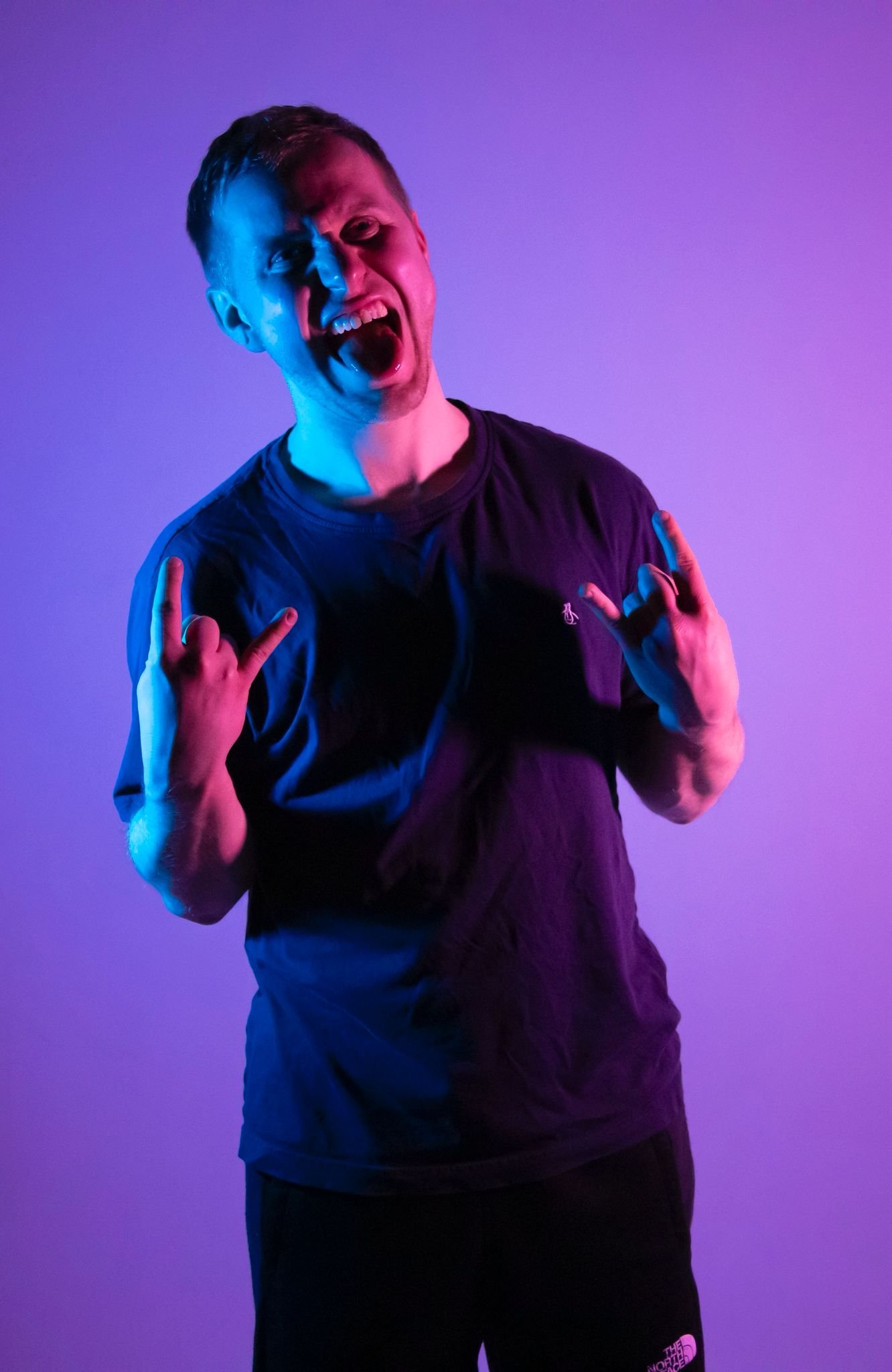 Young man with short hair making a rock and roll gesture with both hands, smiling, wearing a black t-shirt, against a purple background with colorful lighting.