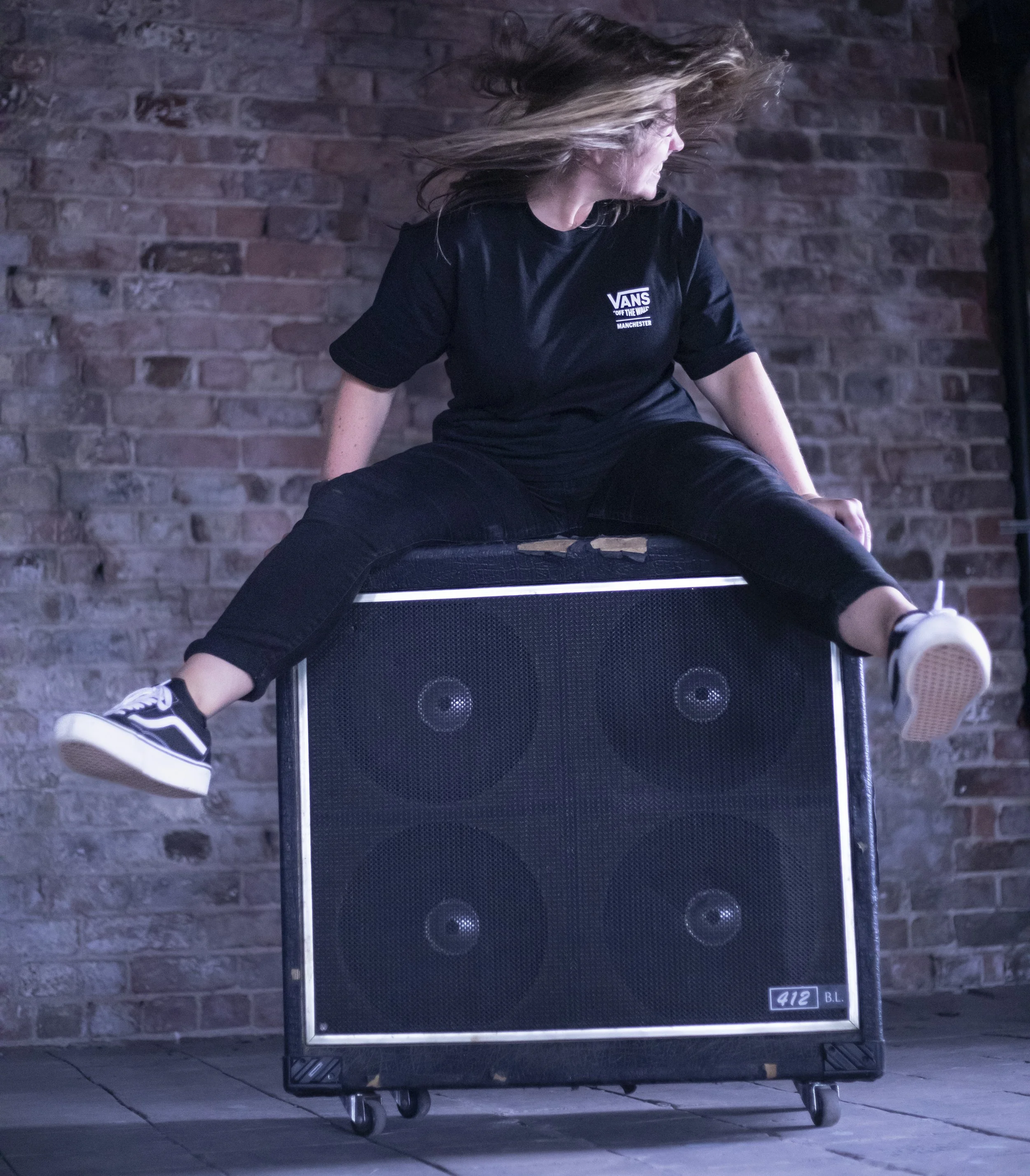 A person sitting on top of a large black speaker cabinet with four speakers, in front of a brick wall. The person has long hair and is wearing a black T-shirt, black pants, and Vans sneakers, with their hair flying as if moving or dancing.