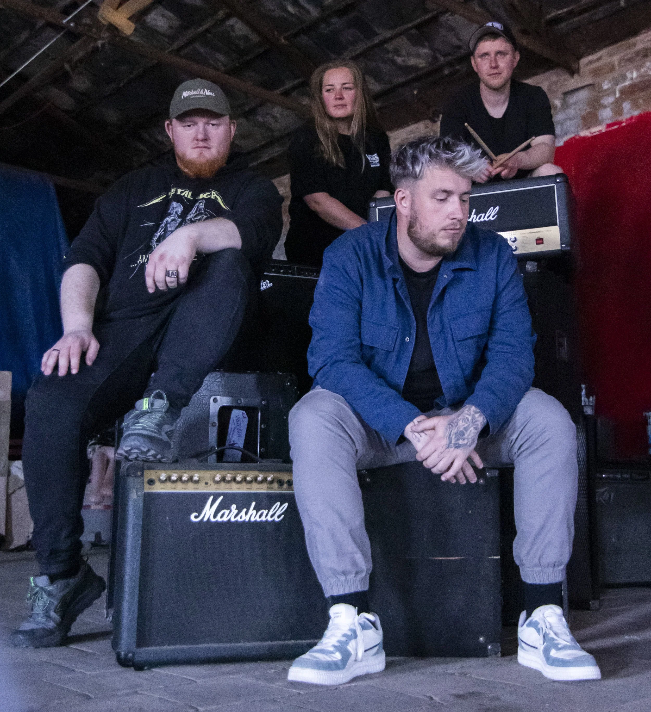 Four people in a music setup with Marshall amplifiers, two men sitting in front and two women behind, in an indoor space with an unfinished ceiling.