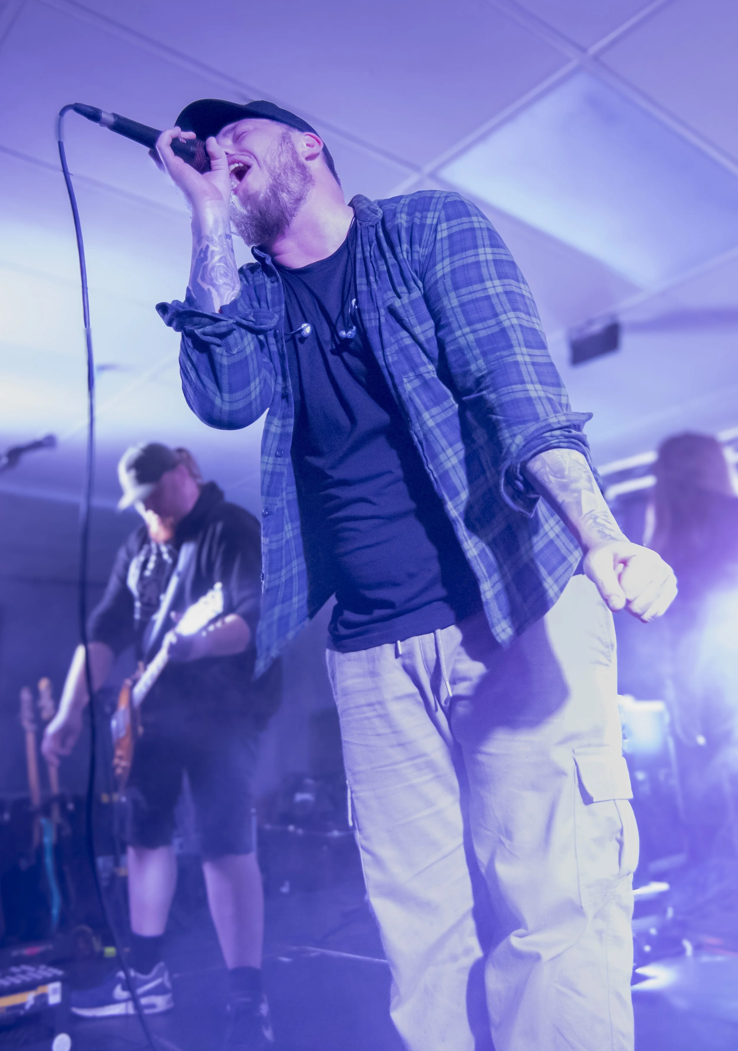 A male singer passionately performing on stage with a microphone, wearing a baseball cap, plaid shirt, and beige cargo pants, while a guitarist plays in the background. The scene is lit with purple and blue stage lights.