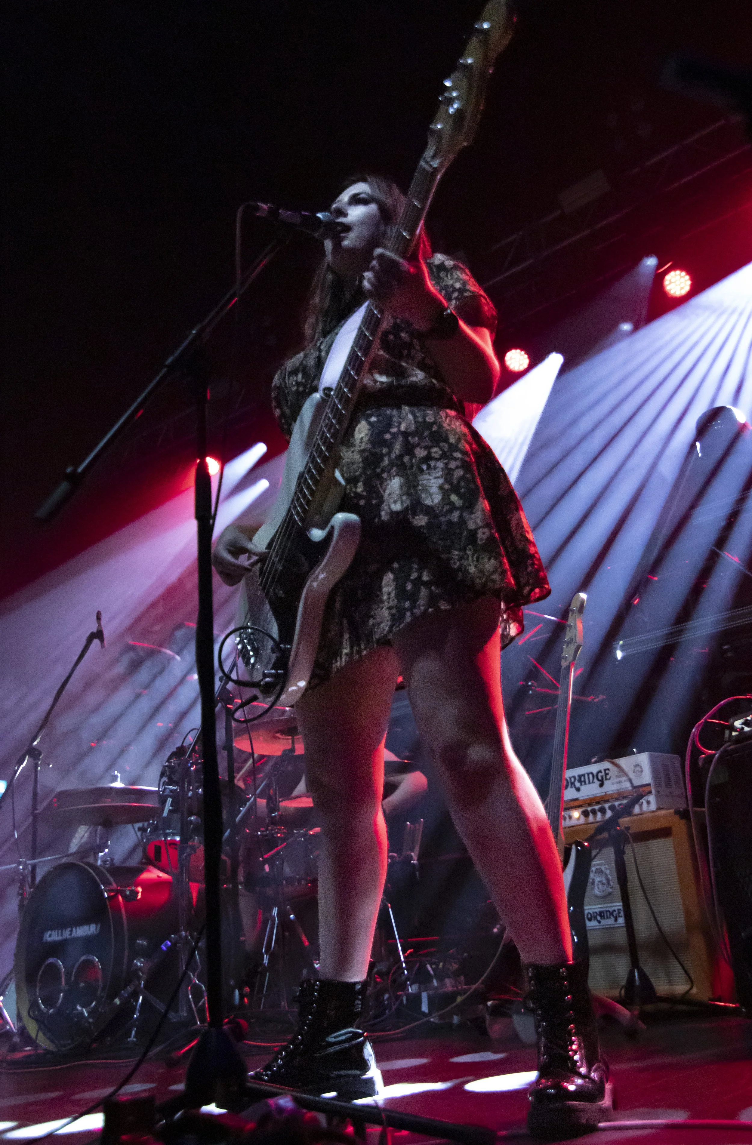 Female musician performing on stage with an electric guitar, wearing a floral dress, black combat boots, and singing into a microphone, with stage lights and musical equipment in the background.