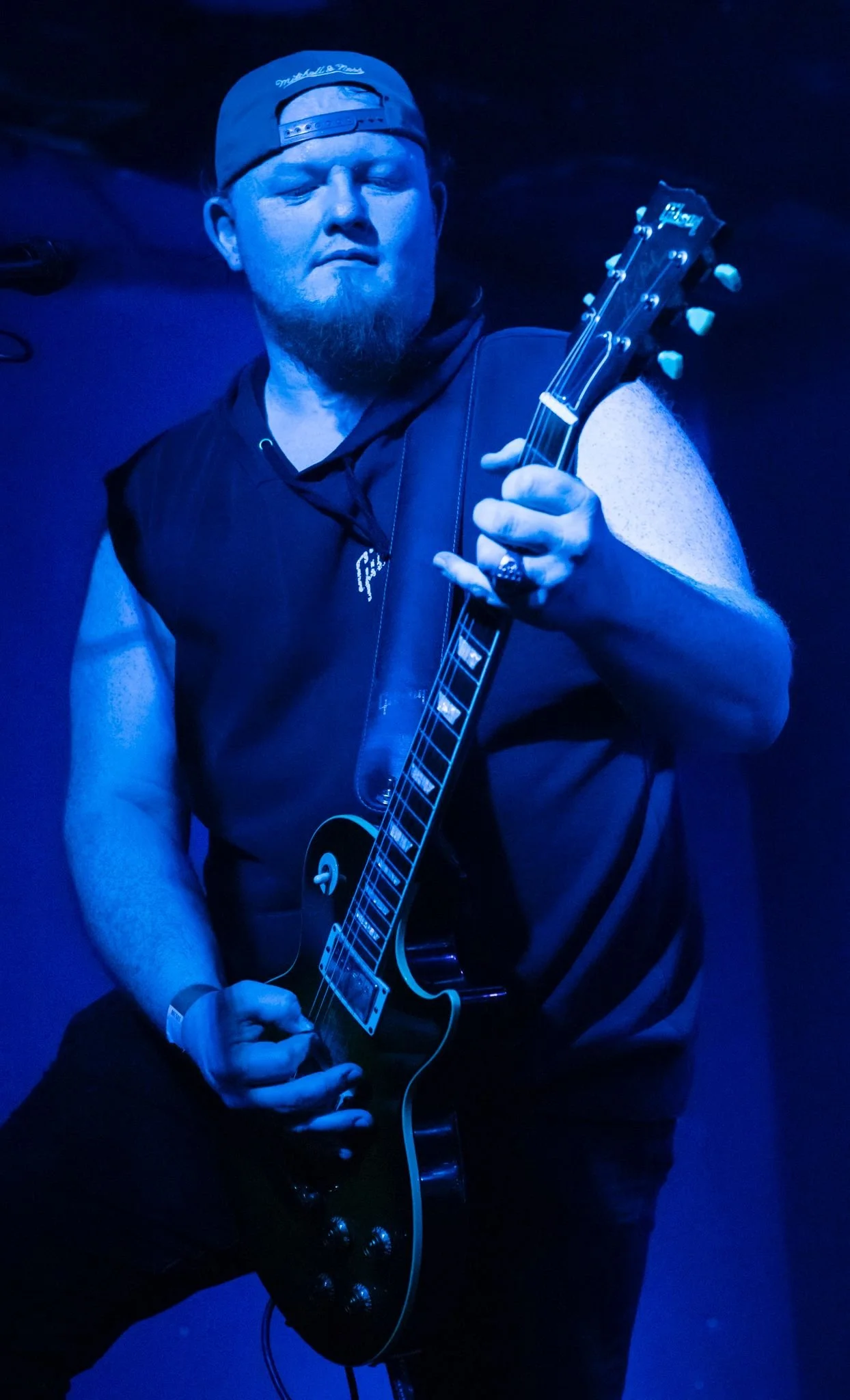 A male guitarist performing on stage under blue lighting, wearing a backwards cap and sleeveless hoodie.