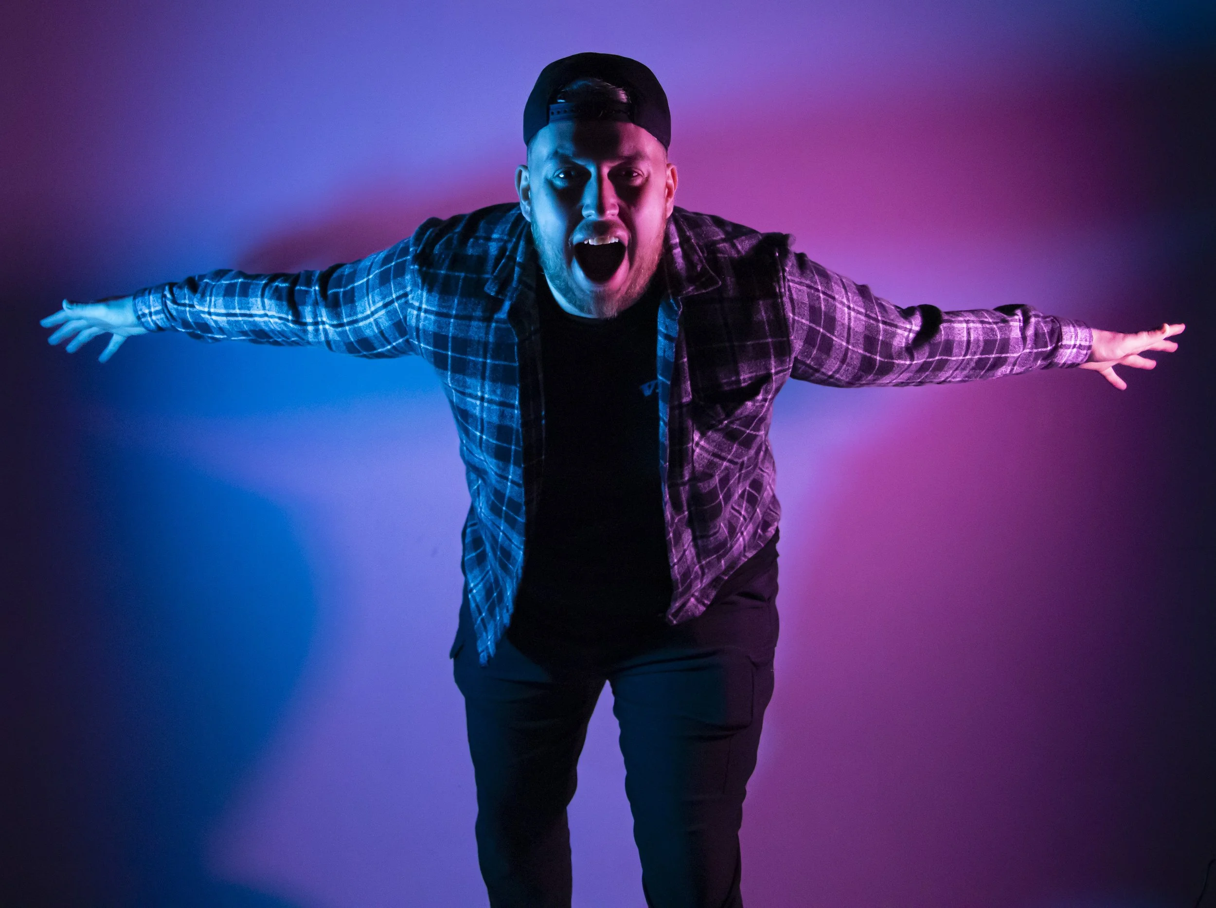 A man wearing a black cap and a plaid shirt with arms outstretched, standing against a colorful gradient background, with his mouth open as if shouting or singing.