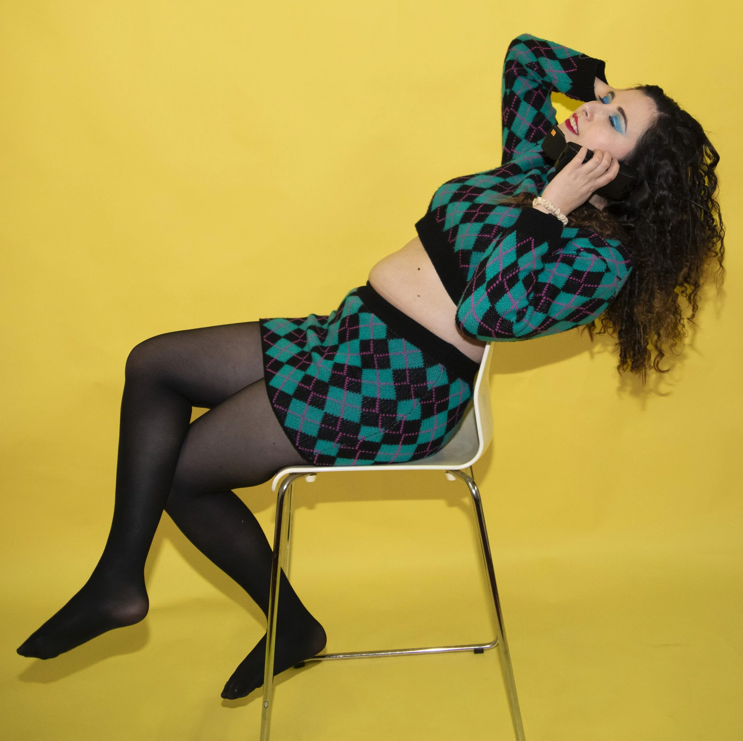A woman with curly hair wearing a matching teal and black plaid outfit, black tights, and black shoes, sitting on a white chair against a yellow background, talking on a black telephone.