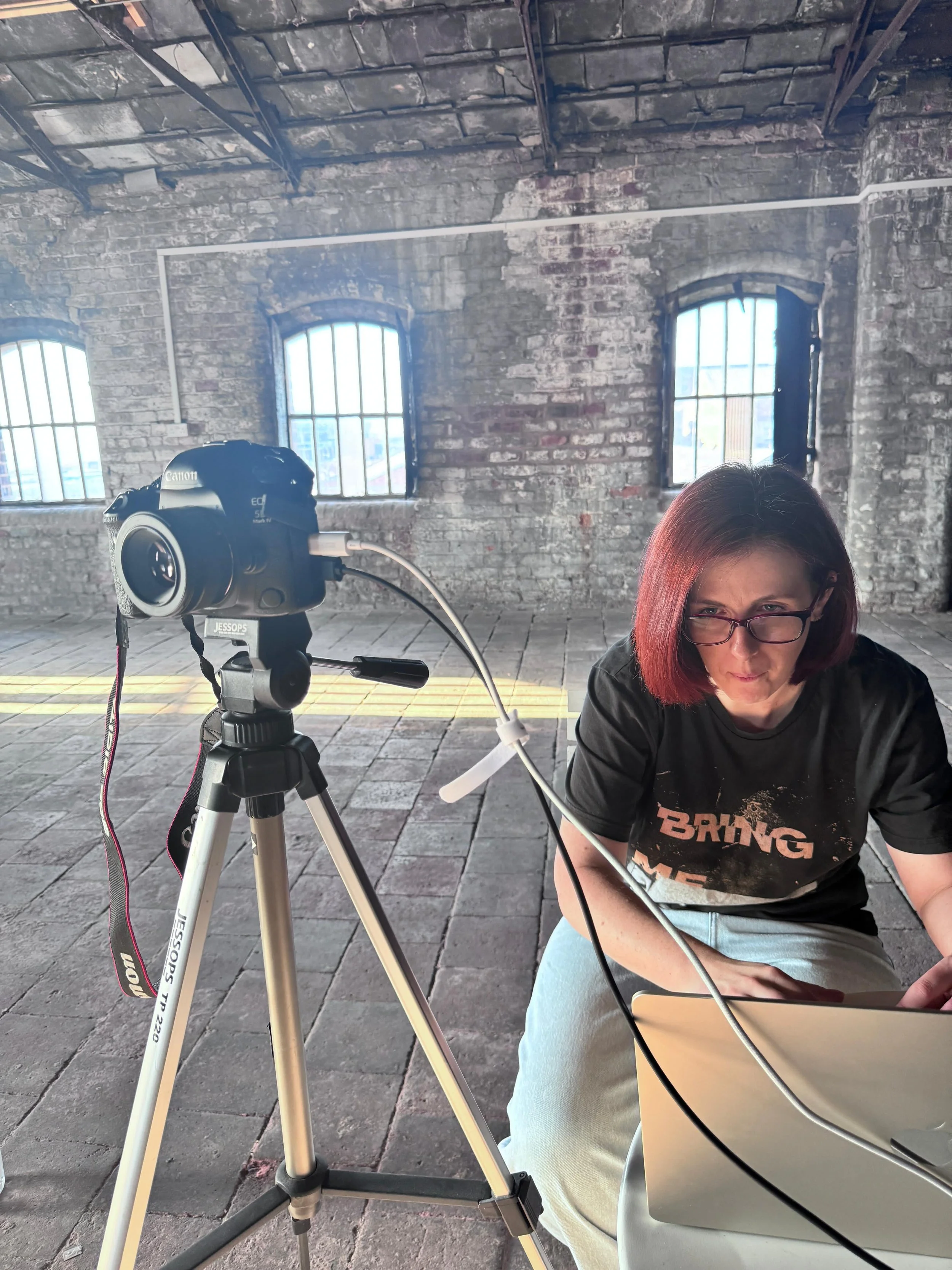 A woman with red hair, glasses, and a black T-shirt crouches beside a laptop in an industrial-style room with exposed brick walls and three arched windows. A Canon camera on a tripod is set up nearby.