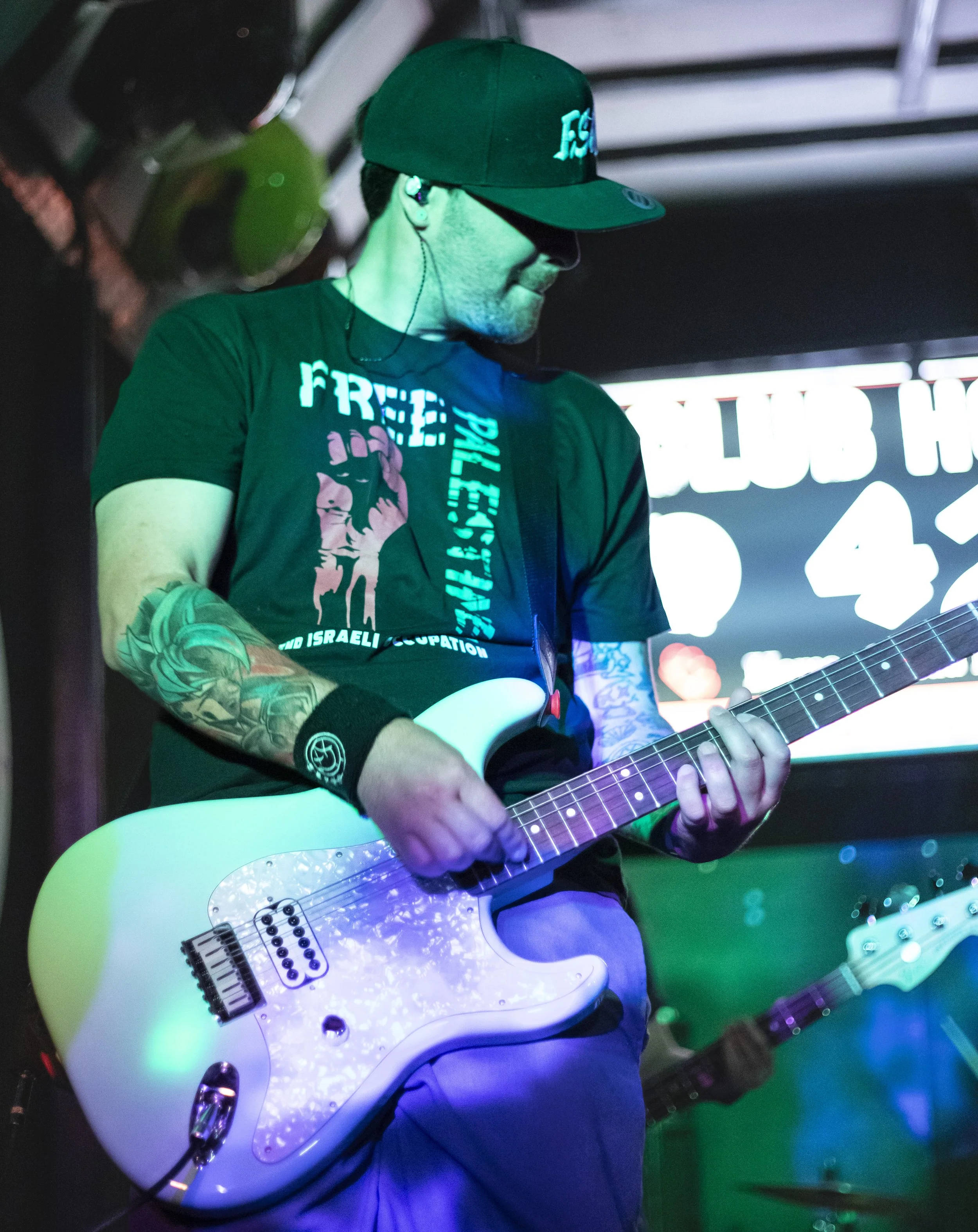 A man playing an electric guitar in a nightclub, wearing a black baseball cap, a black T-shirt with printed text and graphics, and tattoos on his arms. The background shows a decorated digital screen and musical equipment.