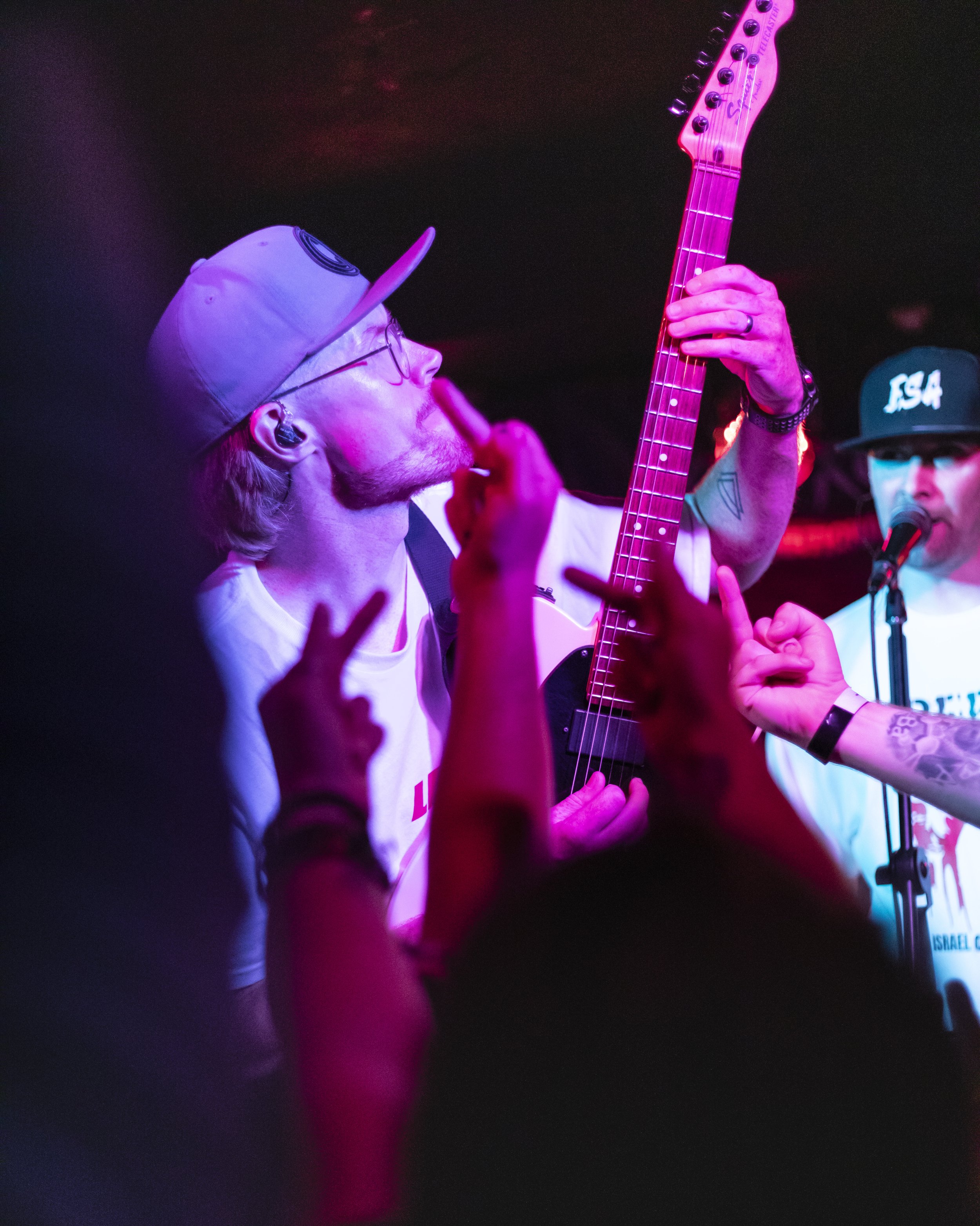 A musician with glasses, a beard, and a baseball cap playing an electric guitar, surrounded by audience members making hand gestures, under colorful stage lights.