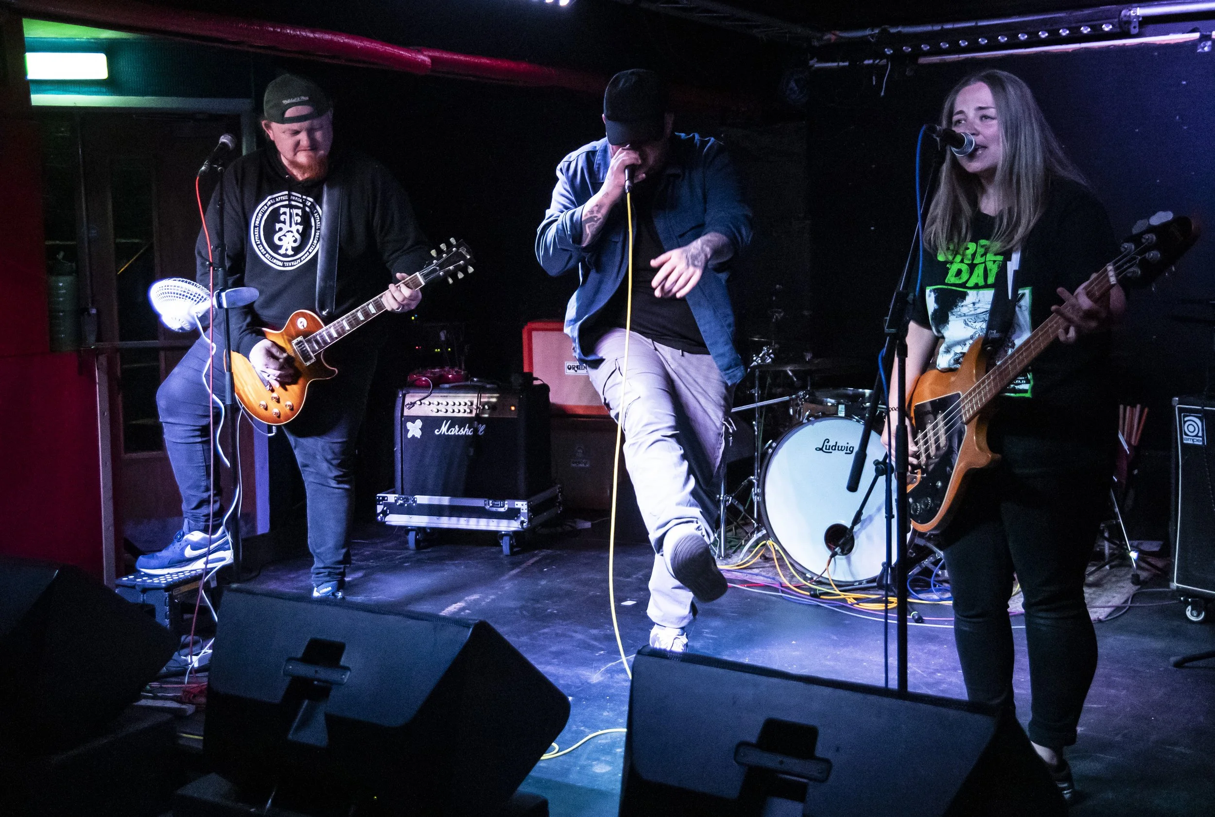 A band performs on stage in a dimly lit venue. The band includes a male guitarist on the left, a male vocalist in the center, and a female bassist on the right. The stage has musical equipment and a drum set in the background.