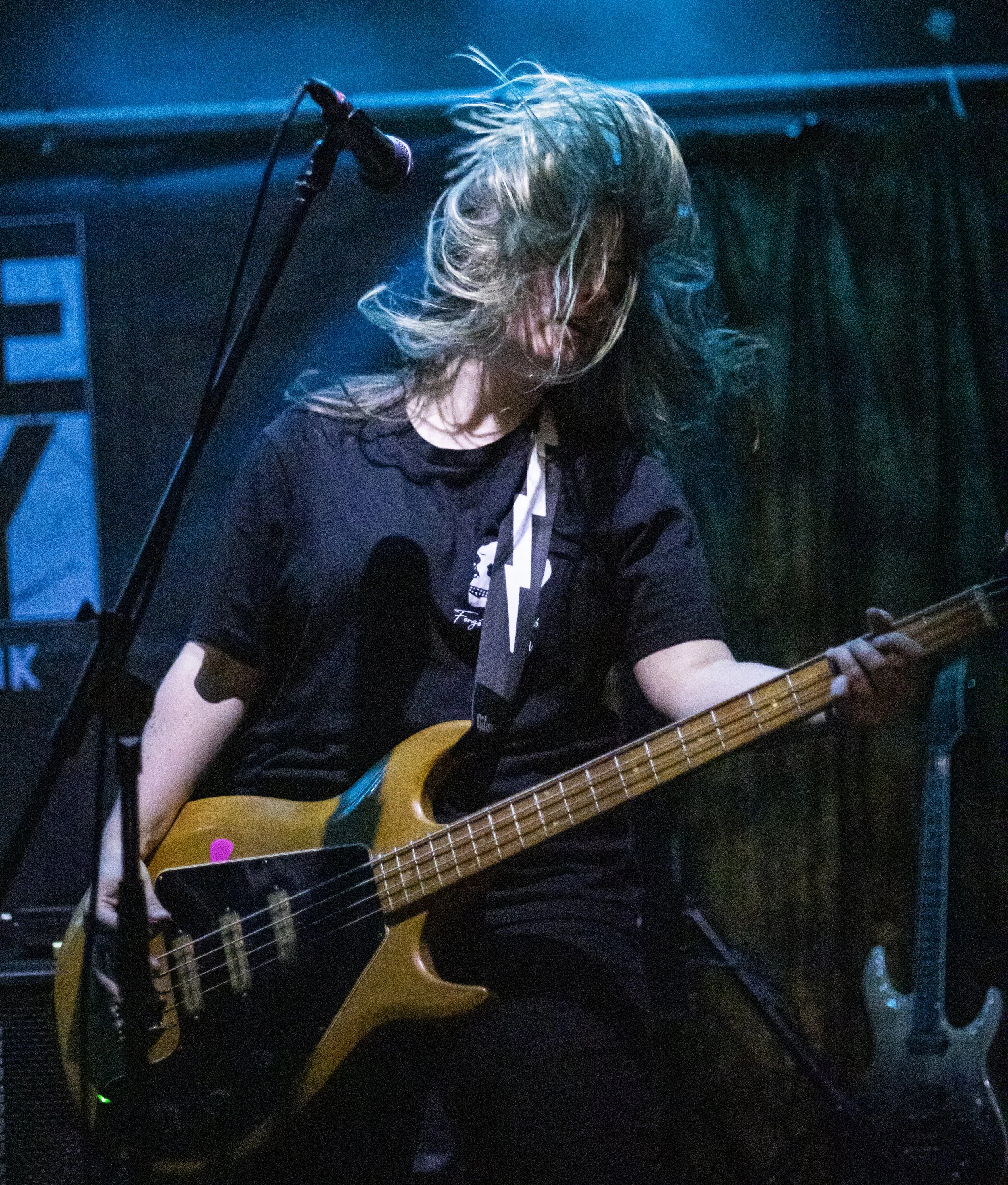 A musician playing an electric bass guitar on stage, with long hair moving as they perform, accompanied by a microphone and stage equipment.