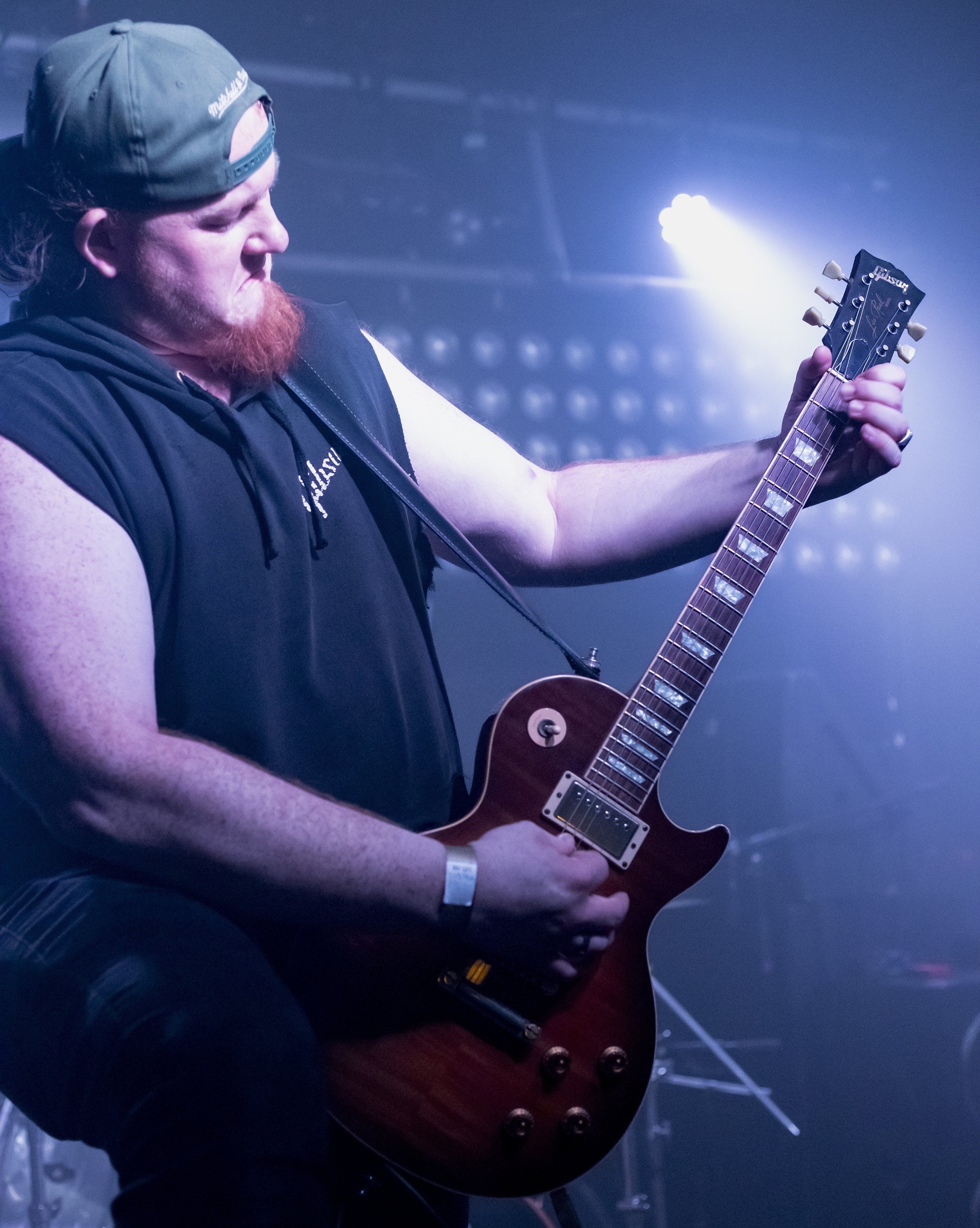 A man with a beard and a backwards cap playing an electric guitar on stage with blue lighting.