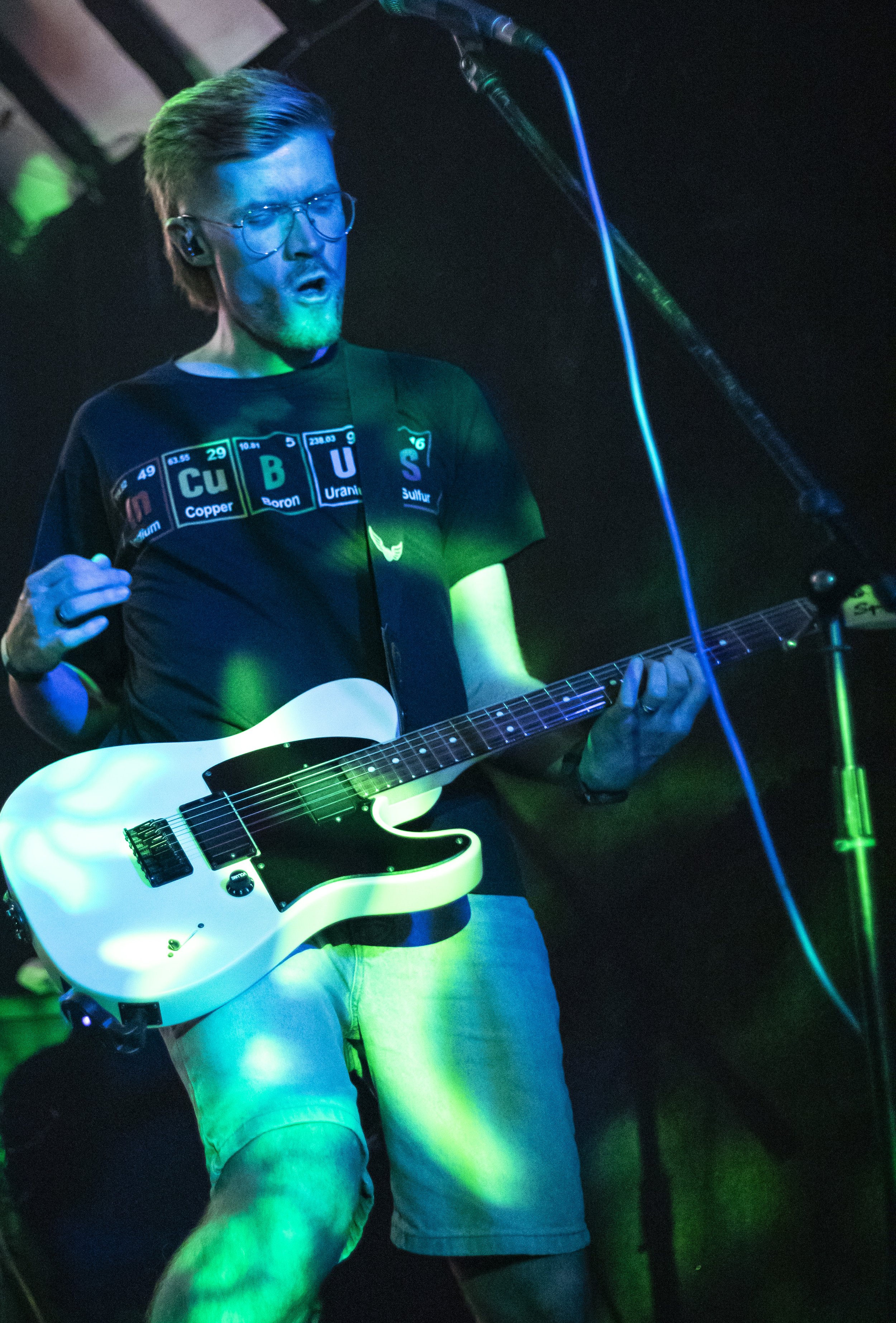 A man with glasses, a beard, and long hair is playing an electric guitar on stage, illuminated by colorful stage lights.