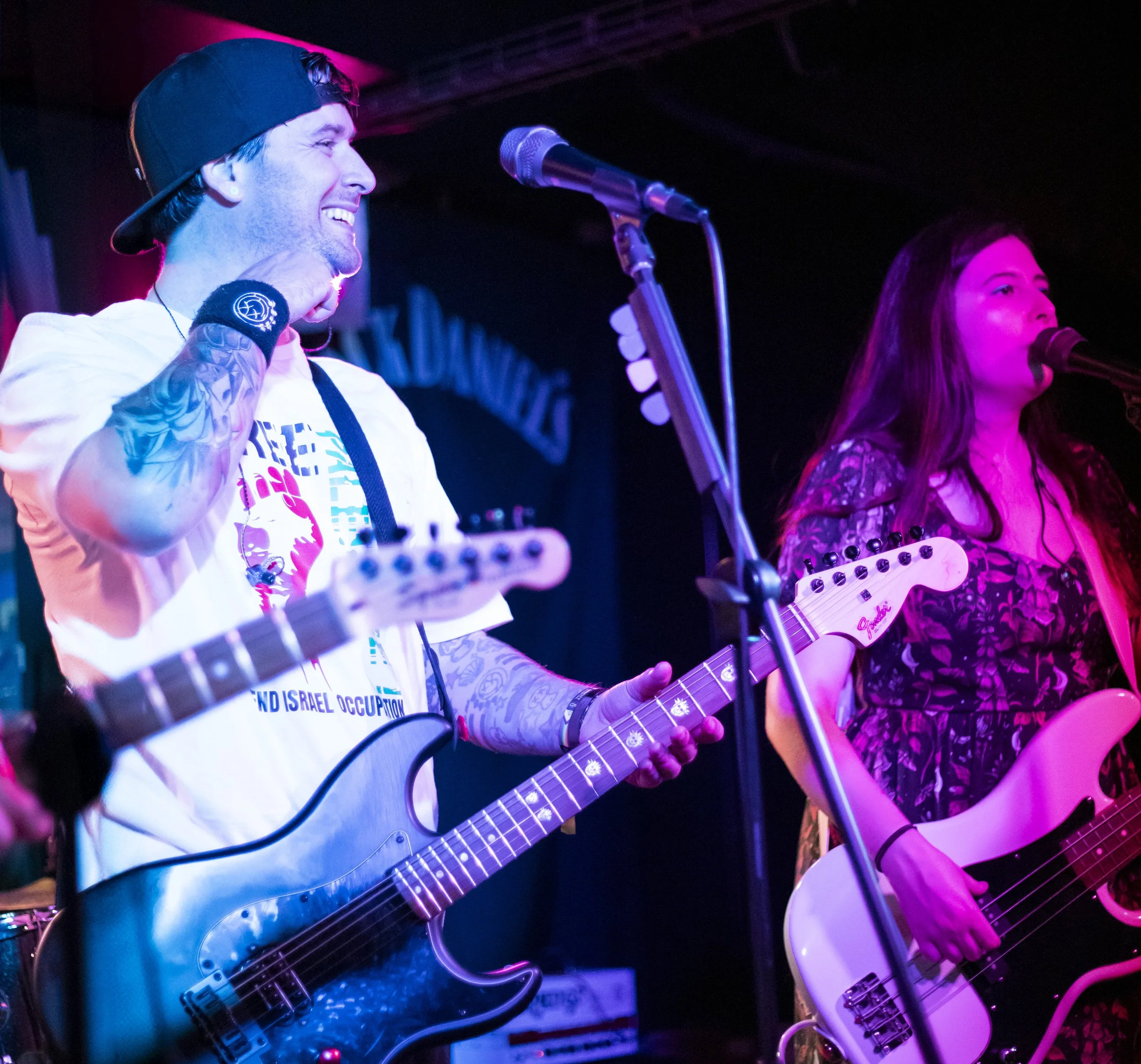 Two musicians performing on stage with guitars, under colorful stage lighting, smiling and singing into microphones.