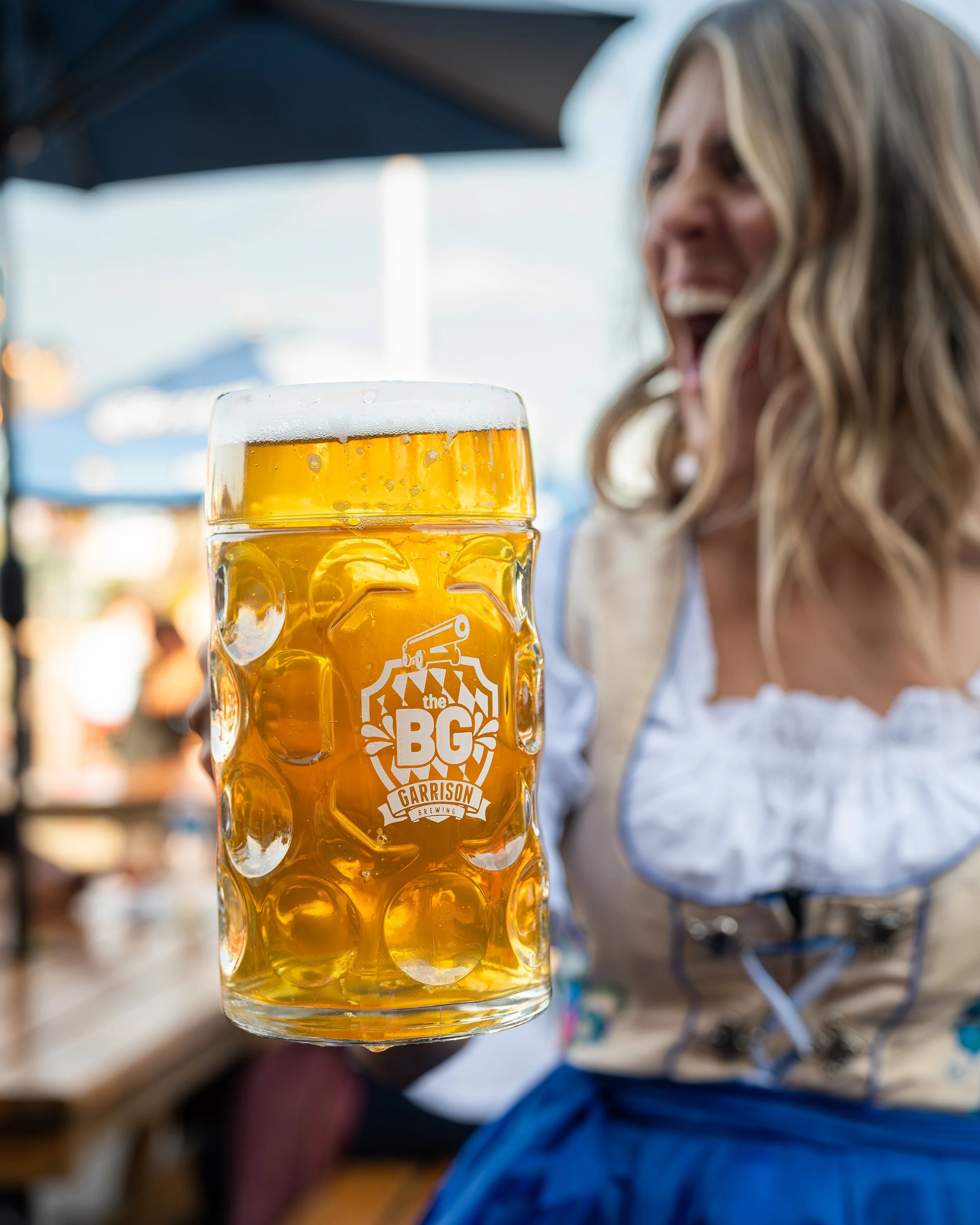 A woman in traditional German attire holding a large glass of beer at an outdoor event, with blurred background.