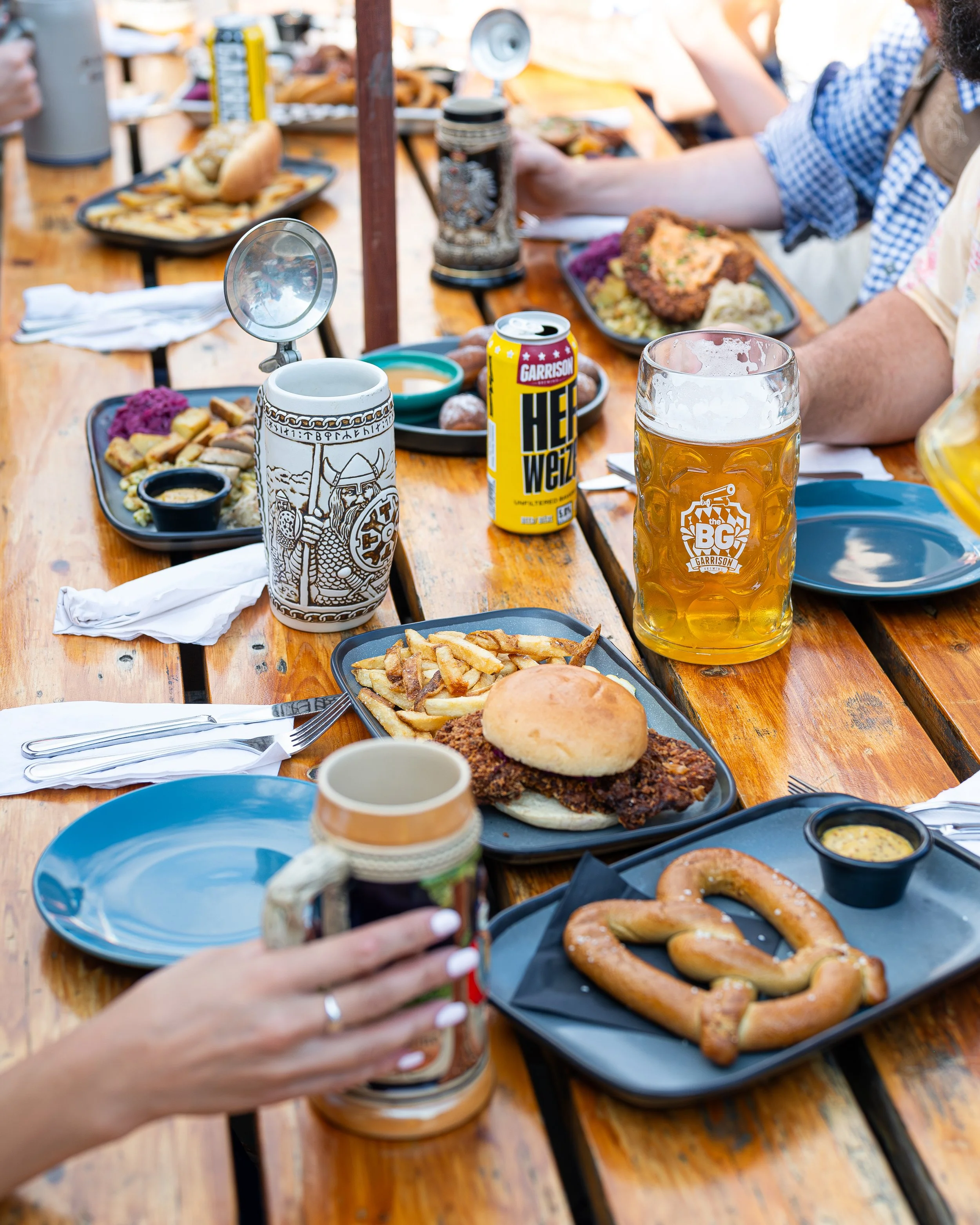 A table with plates of food including fries, a sandwich bun with fried chicken, pretzels with mustard, and various drinks including beer, soda, and a mug. People are gathered around, with some visible hands holding drinks.