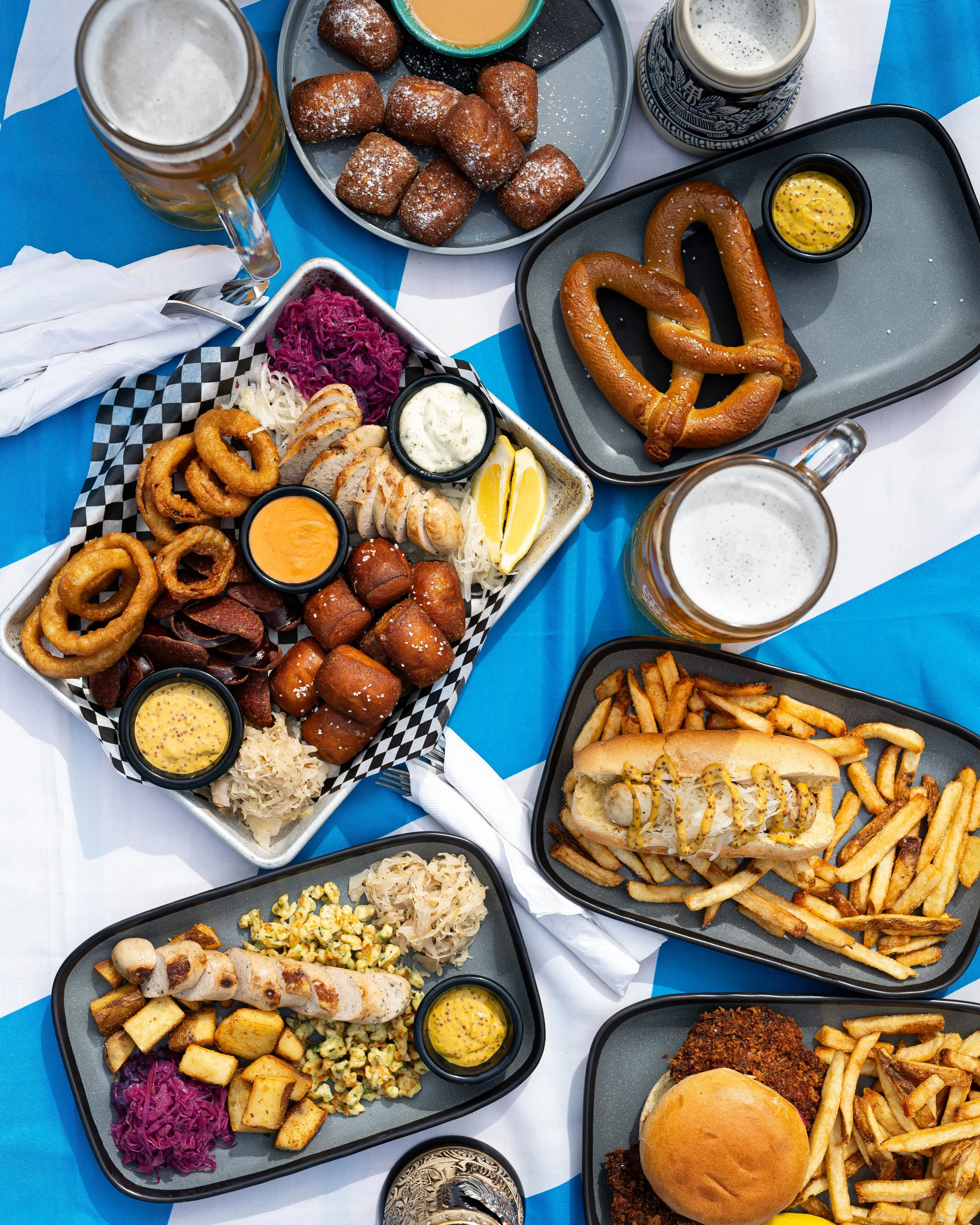 A table with various dishes including fried foods, pretzels, hot dogs, french fries, onion rings, and beverages, likely at an outdoor gathering or festival.