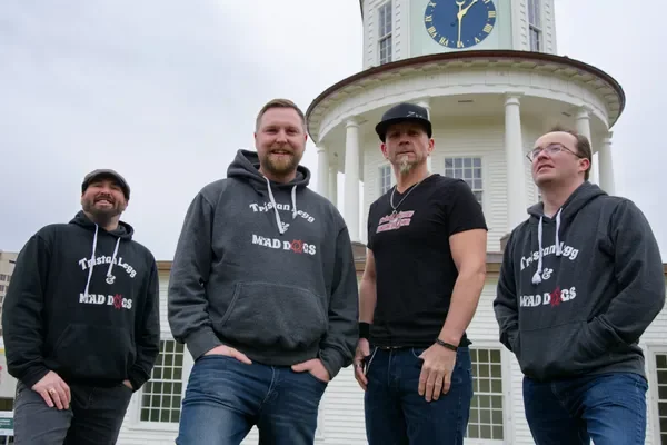 Four men standing outside in front of a white tower with a clock, wearing casual clothing and hoodies with text.