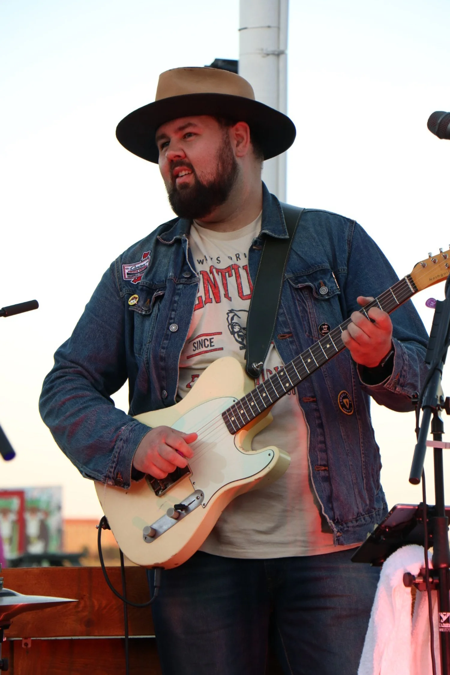 A man wearing a wide-brimmed hat, denim jacket, and a graphic t-shirt playing a white electric guitar on stage at sunset.