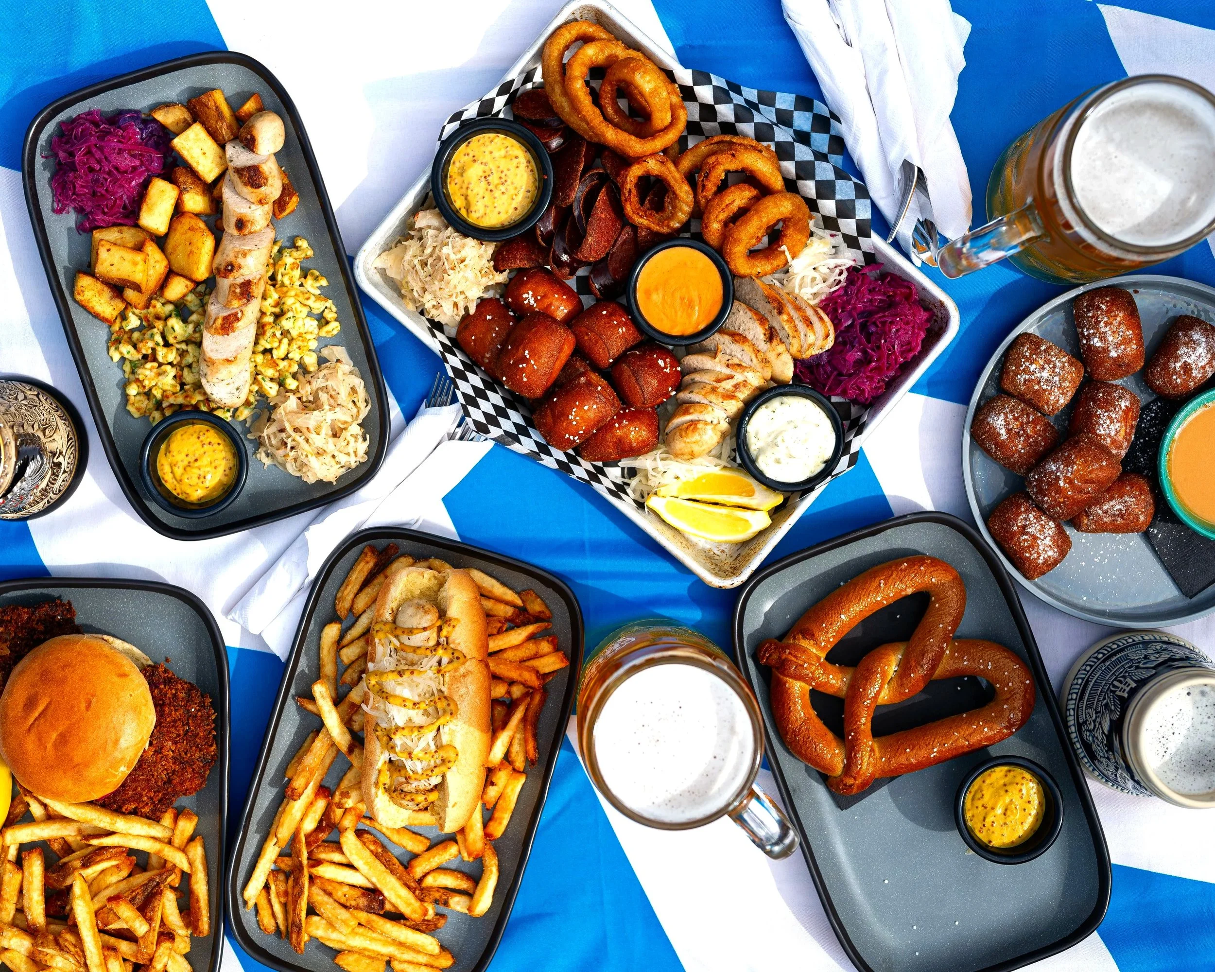A table set with various plates of American bar food, including hot dogs with condiments and fries, onion rings, fried chicken wings, pretzels, fried croquettes, and beverages like beer, with a blue and white tablecloth.