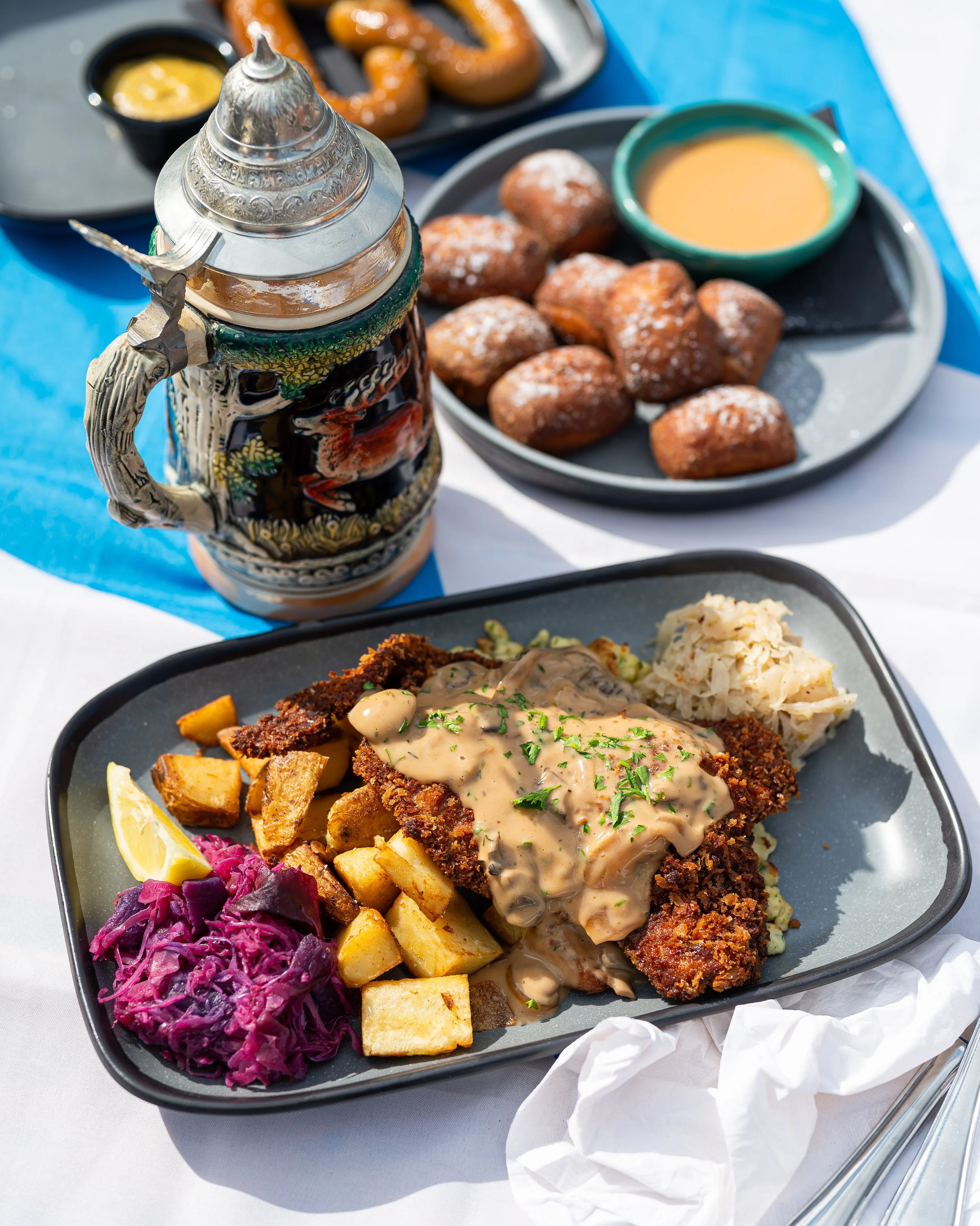 A Battered and fried fish fillet with gravy, served with mashed potatoes, coleslaw, roasted potatoes, lemon wedge, and purple pickled cabbage on a black plate. In the background, there are hush puppies with dipping sauce, a traditional stein of beer,