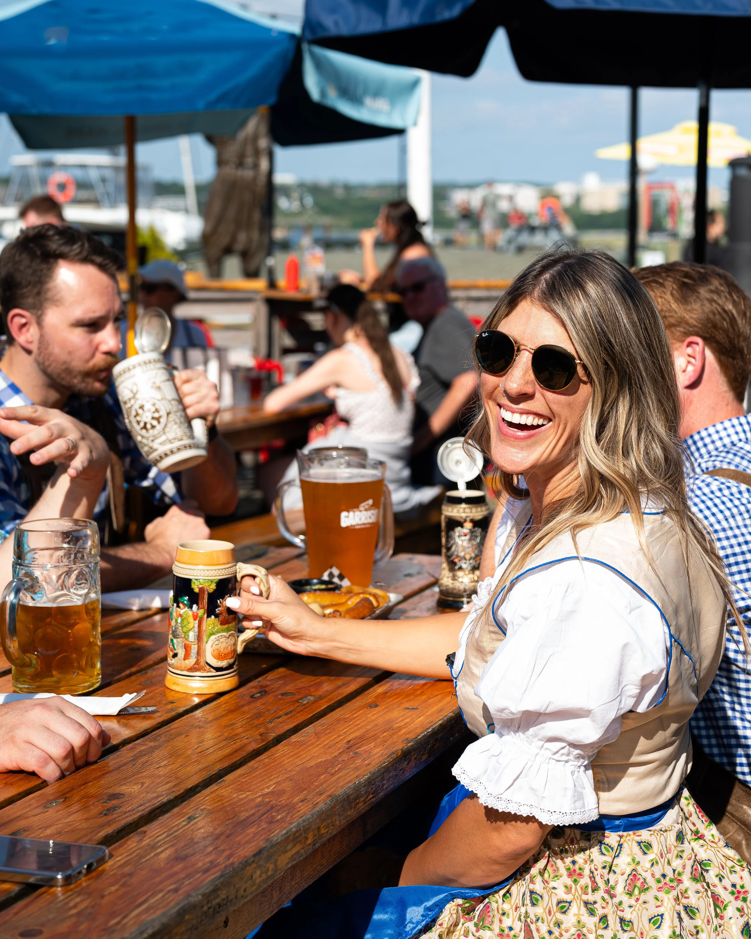 People enjoying drinks and socializing outdoors at a wooden table under umbrellas, with some in traditional or casual summer attire, and a woman smiling and wearing sunglasses.