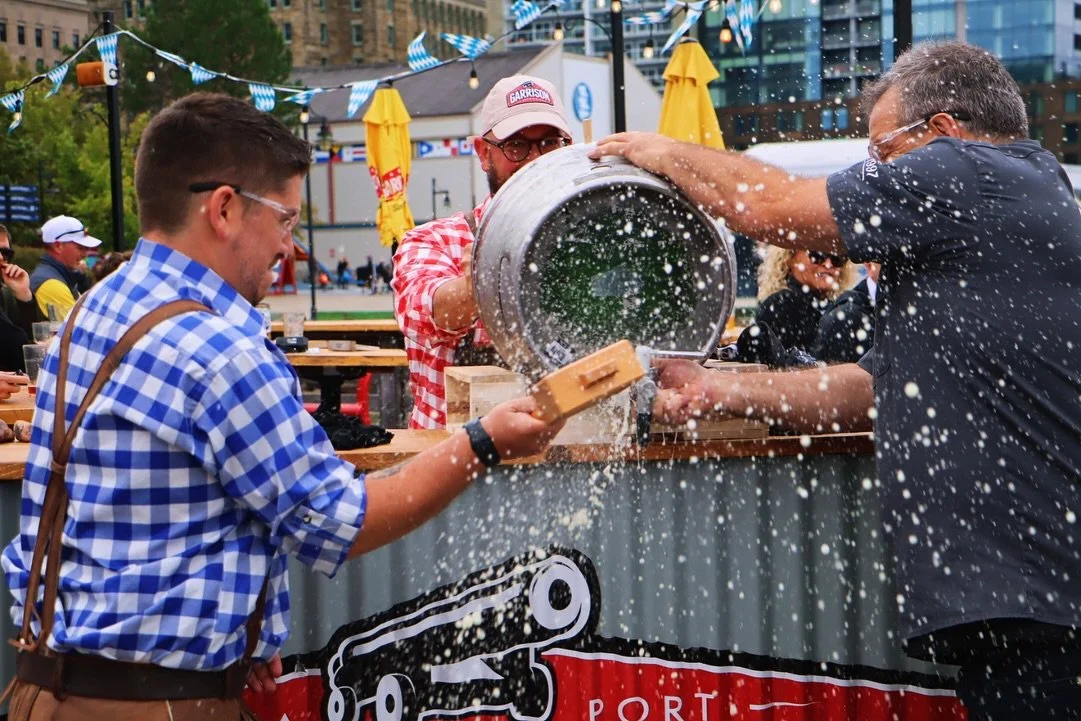 People at a festival pouring beer from a barrel, with foam spilling out, surrounded by colorful decorations and outdoor setting.