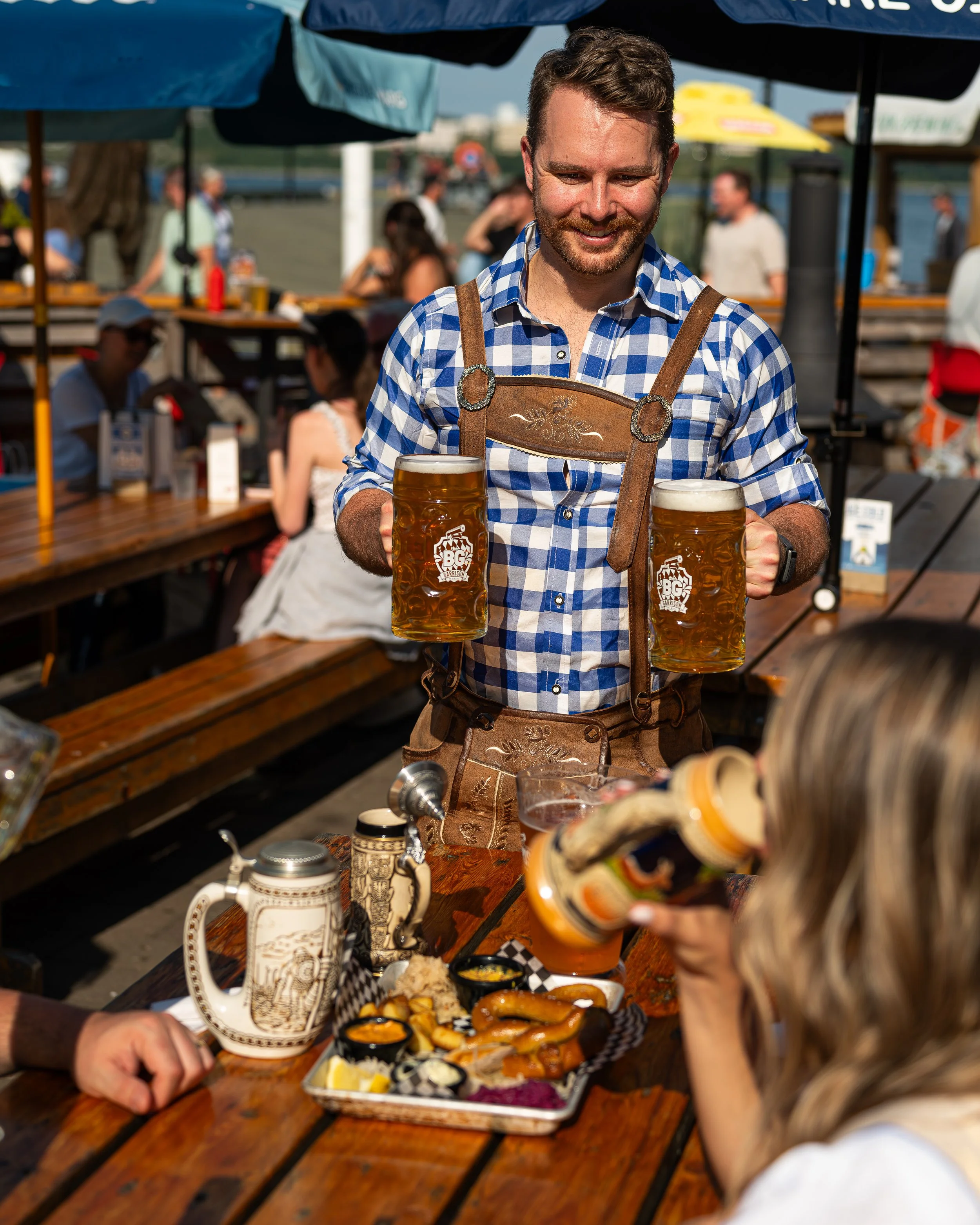 A man in traditional German attire serving beer at an outdoor event, with a table of food and people in the background.