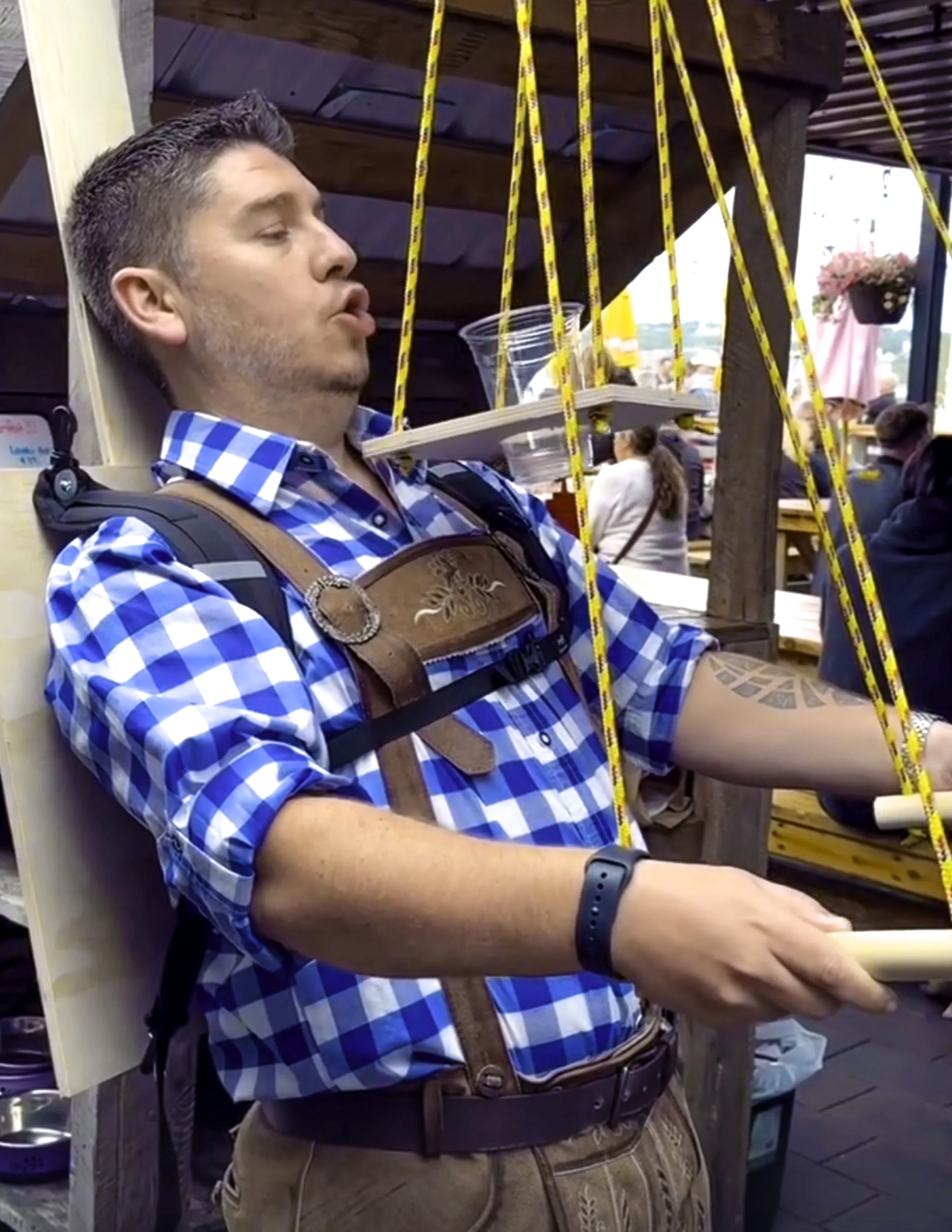 A man in a blue checkered shirt with a backpack and a tattoo on his left arm, holding a tray with a plastic cup on his shoulder, appears to be asleep at a festival or outdoor event with wooden structures and people in the background.