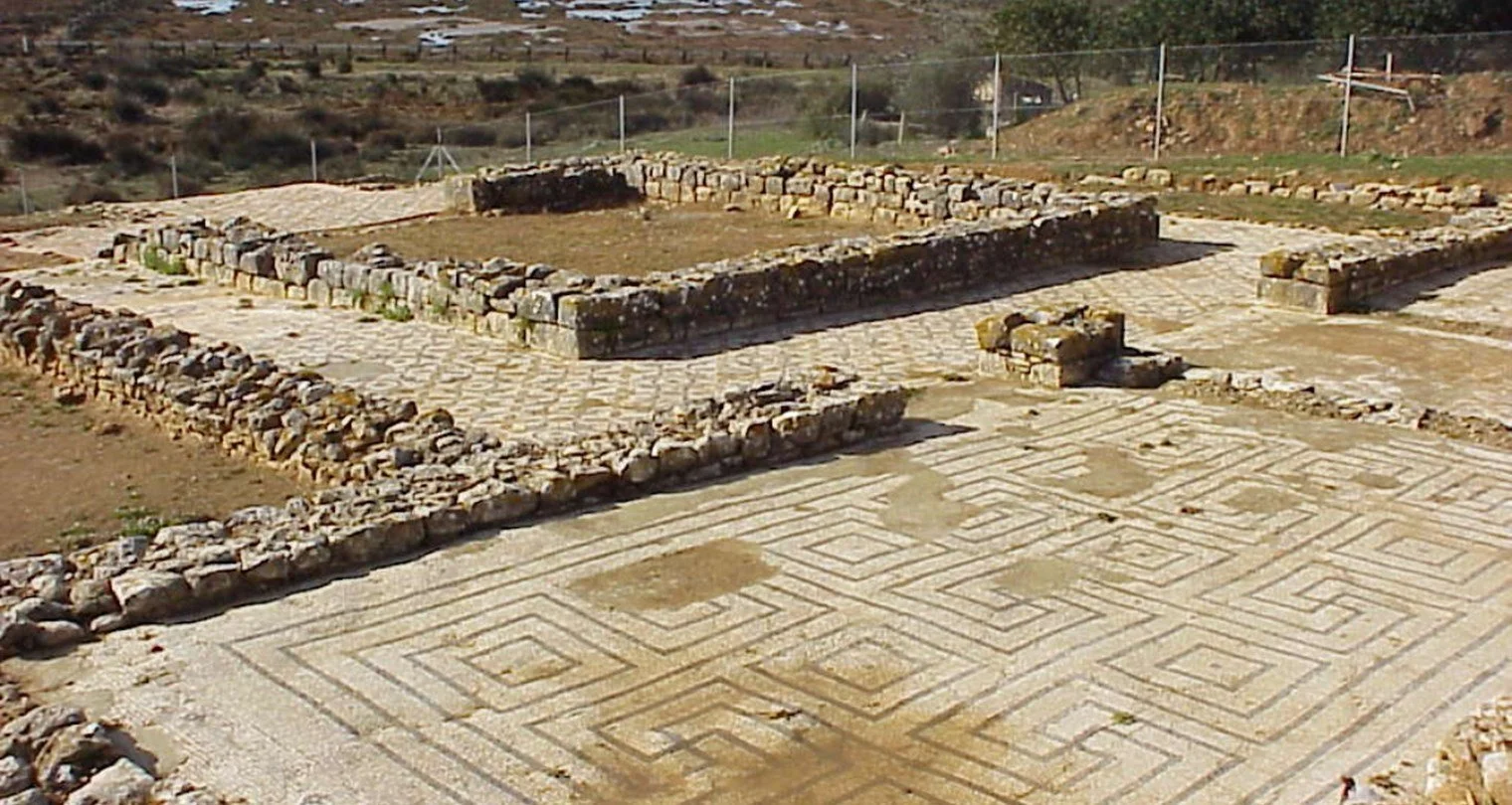 Excavated archaeological site with ancient stone walls and a patterned stone floor, surrounded by a fence in Alvor