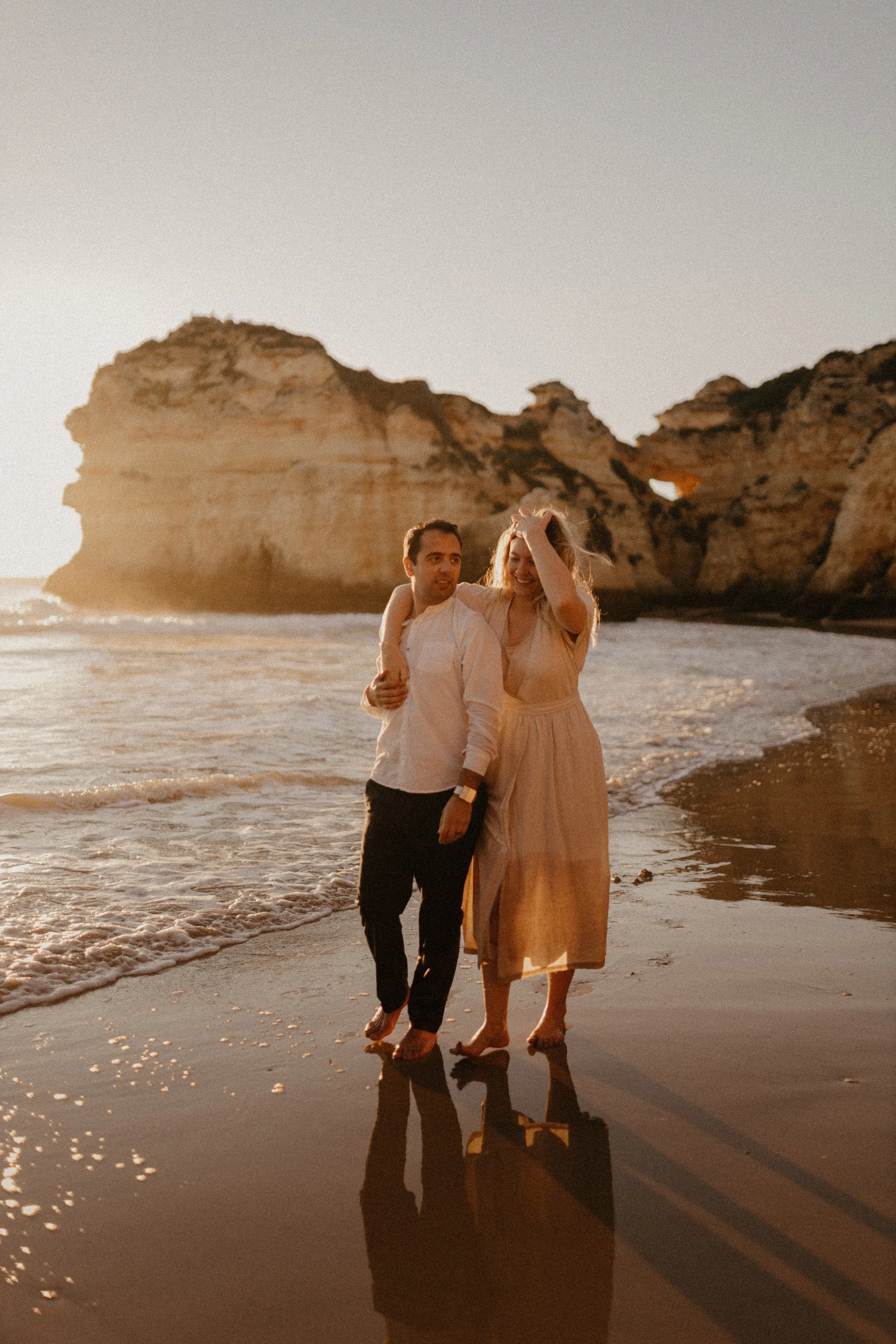 A couple walking along the beach with cliffs in the background during sunset. Discover Alvor - best spots to eat, drink, and experience on your holiday.