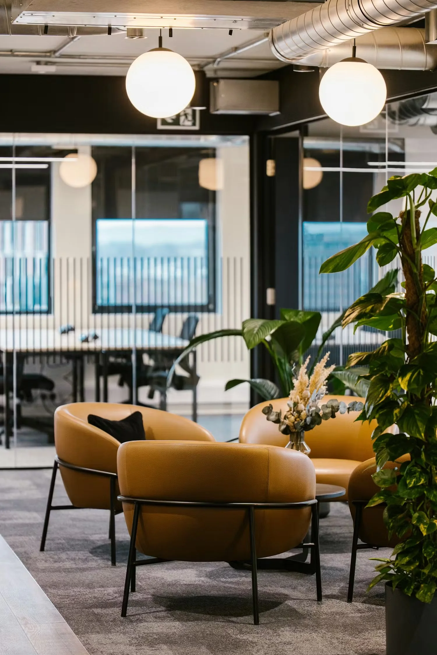 Modern office lounge area with mustard-colored chairs, a glass table with flowers, and large green plants, with a meeting room in the background.