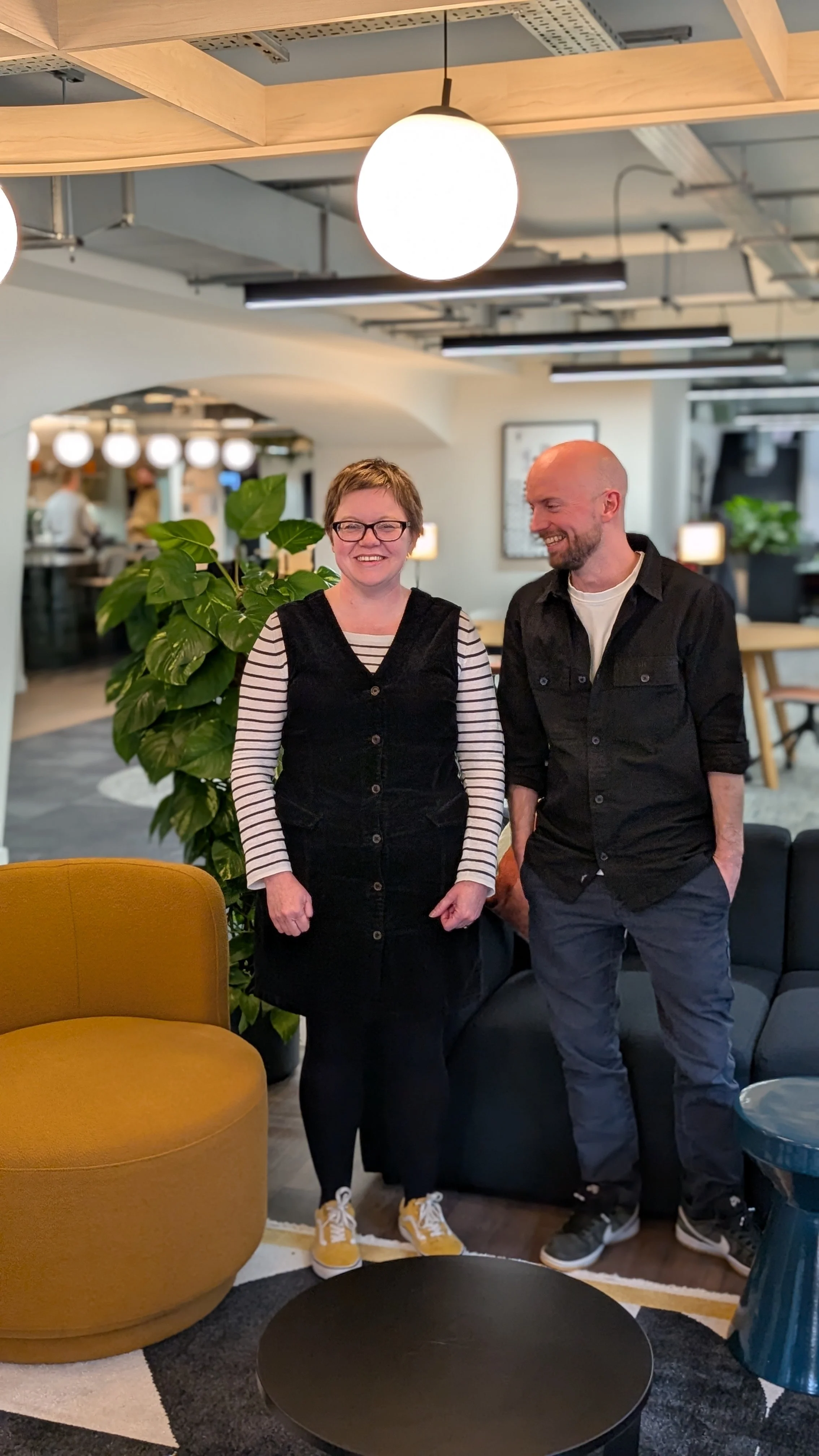 A woman and a man standing together in a modern, well-lit indoor setting, smiling, with a large green plant behind them, a mustard-colored chair to the left, and a black sofa to the right.