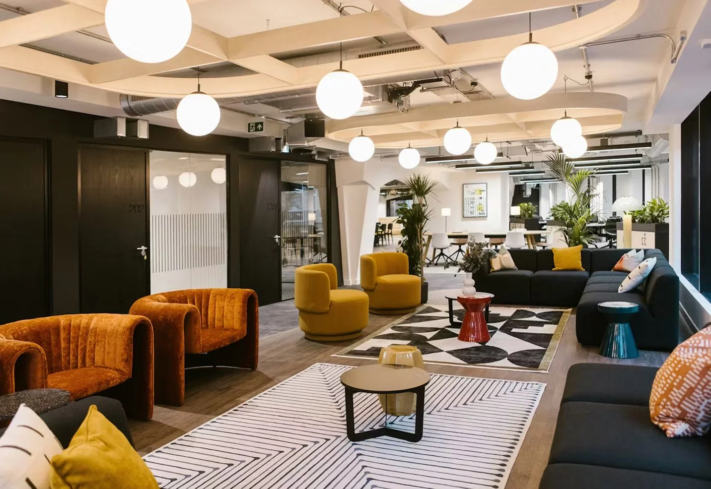 Modern lobby area with colorful chairs and sofas, decorative plants, patterned rugs, and hanging spherical ceiling lights.