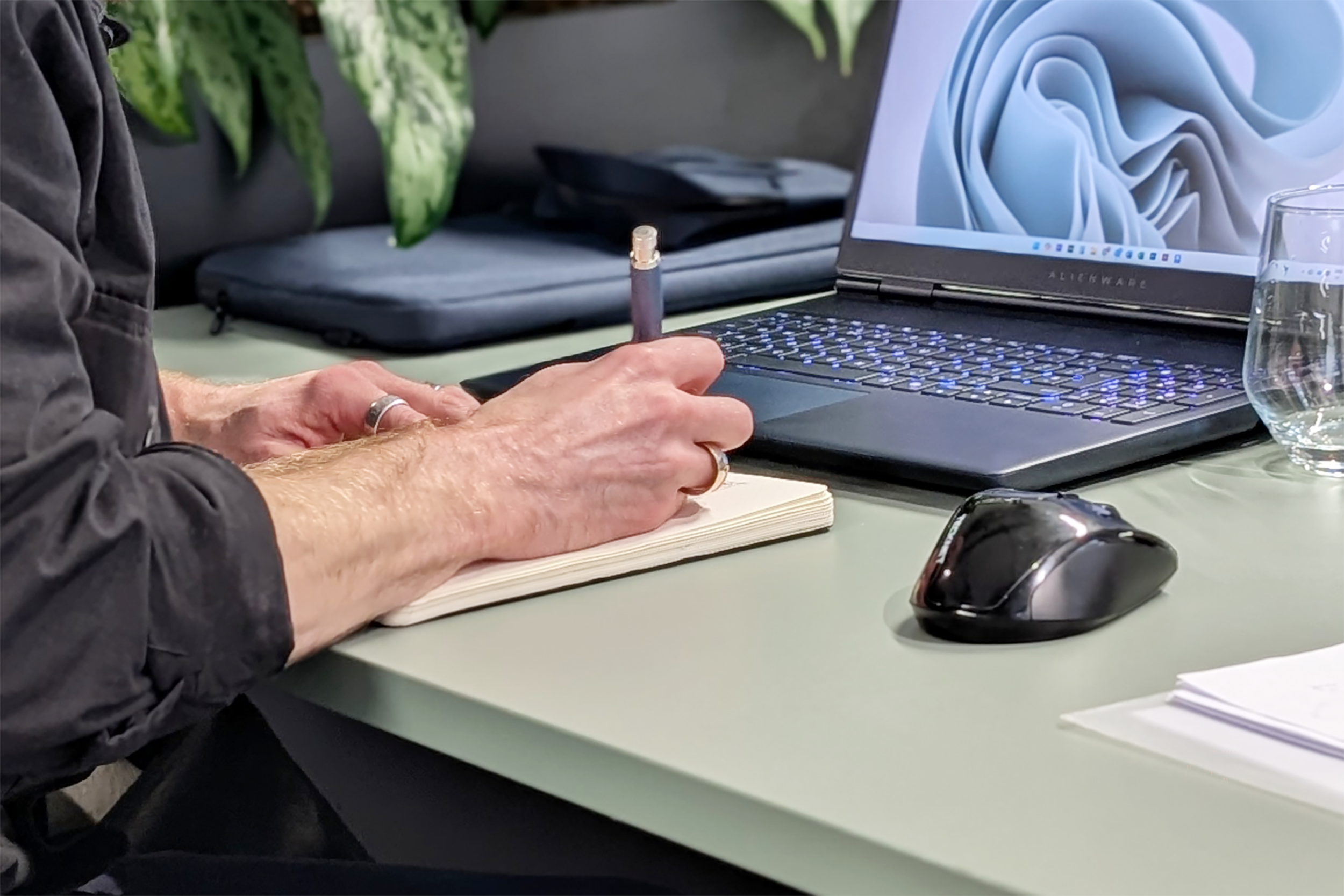 Person writing in notebook with laptop, mouse, glass of water, and documents on desk.