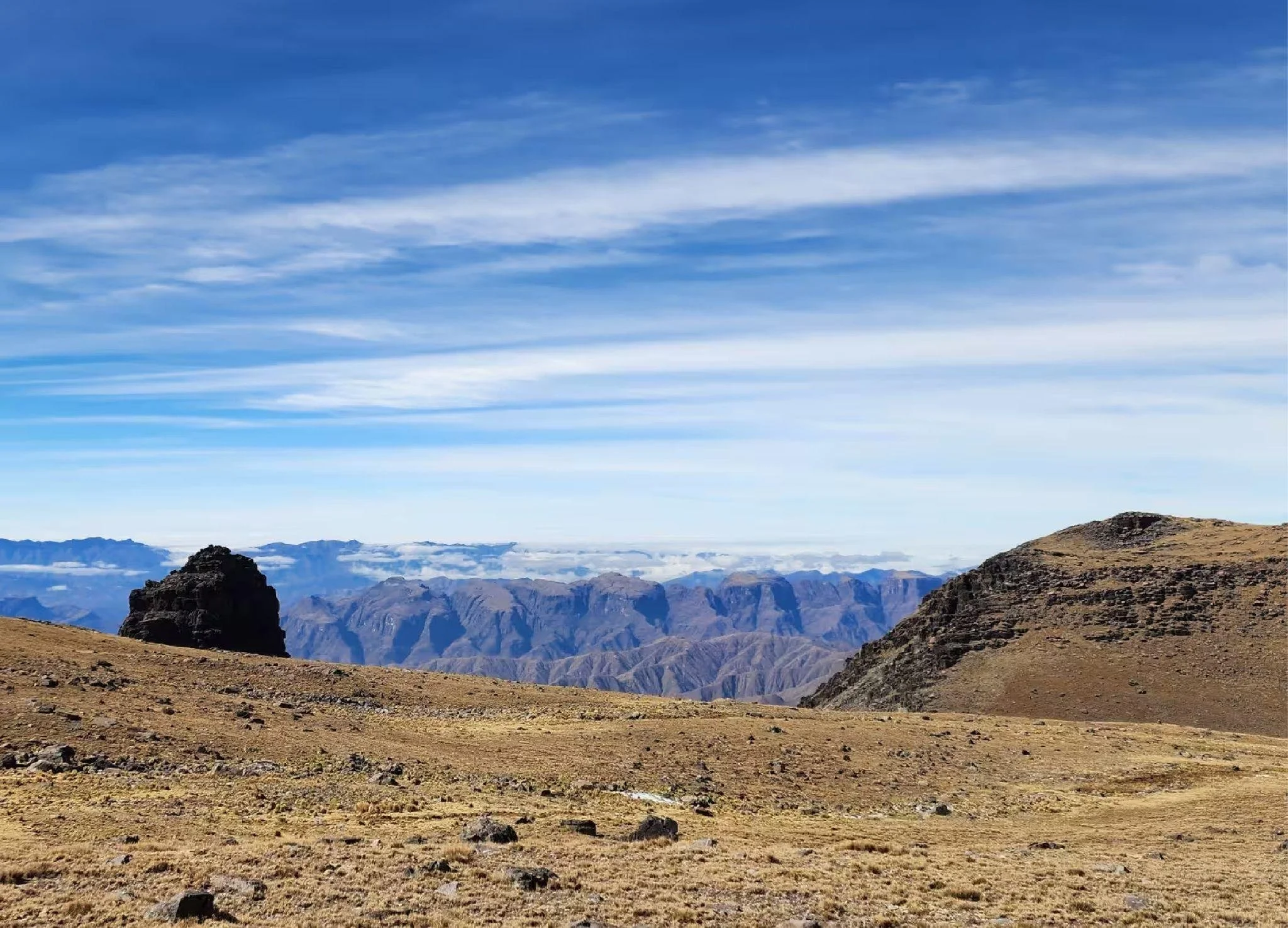 Berglandschaft mit trockenem, bewaldetem Gelände im Vordergrund, hohen Bergen im Hintergrund und einer blauen Himmel mit Wolken.