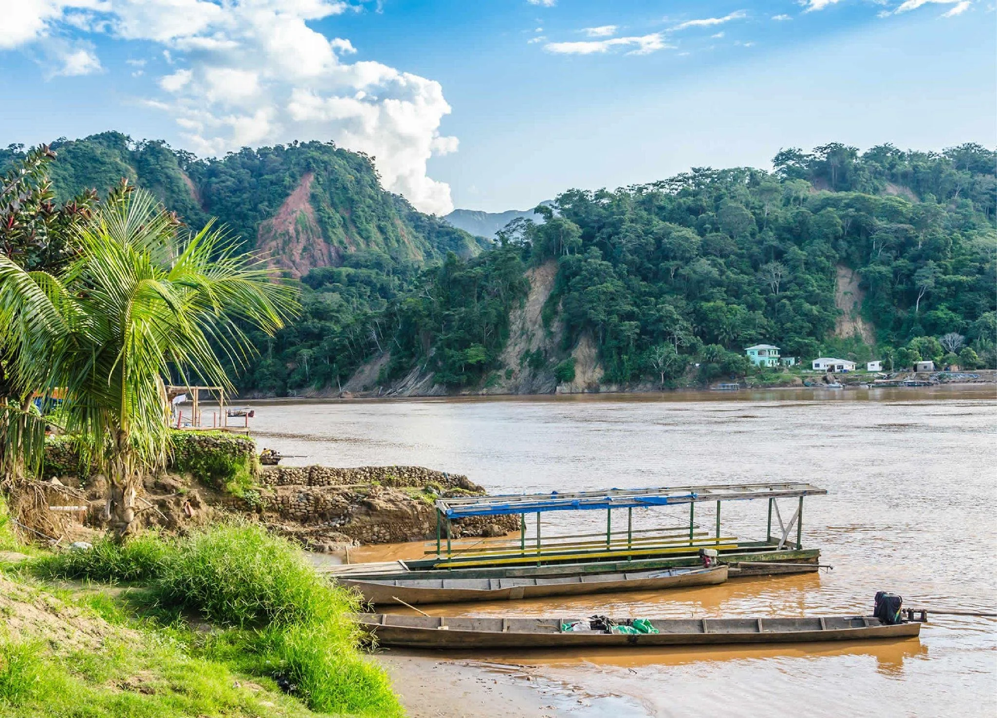 Blick auf einen Fluss mit Booten, grüne Hügel im Hintergrund, am Ufer sind Bäume und kleine Gebäude zu sehen, bei sonnigem Himmel.