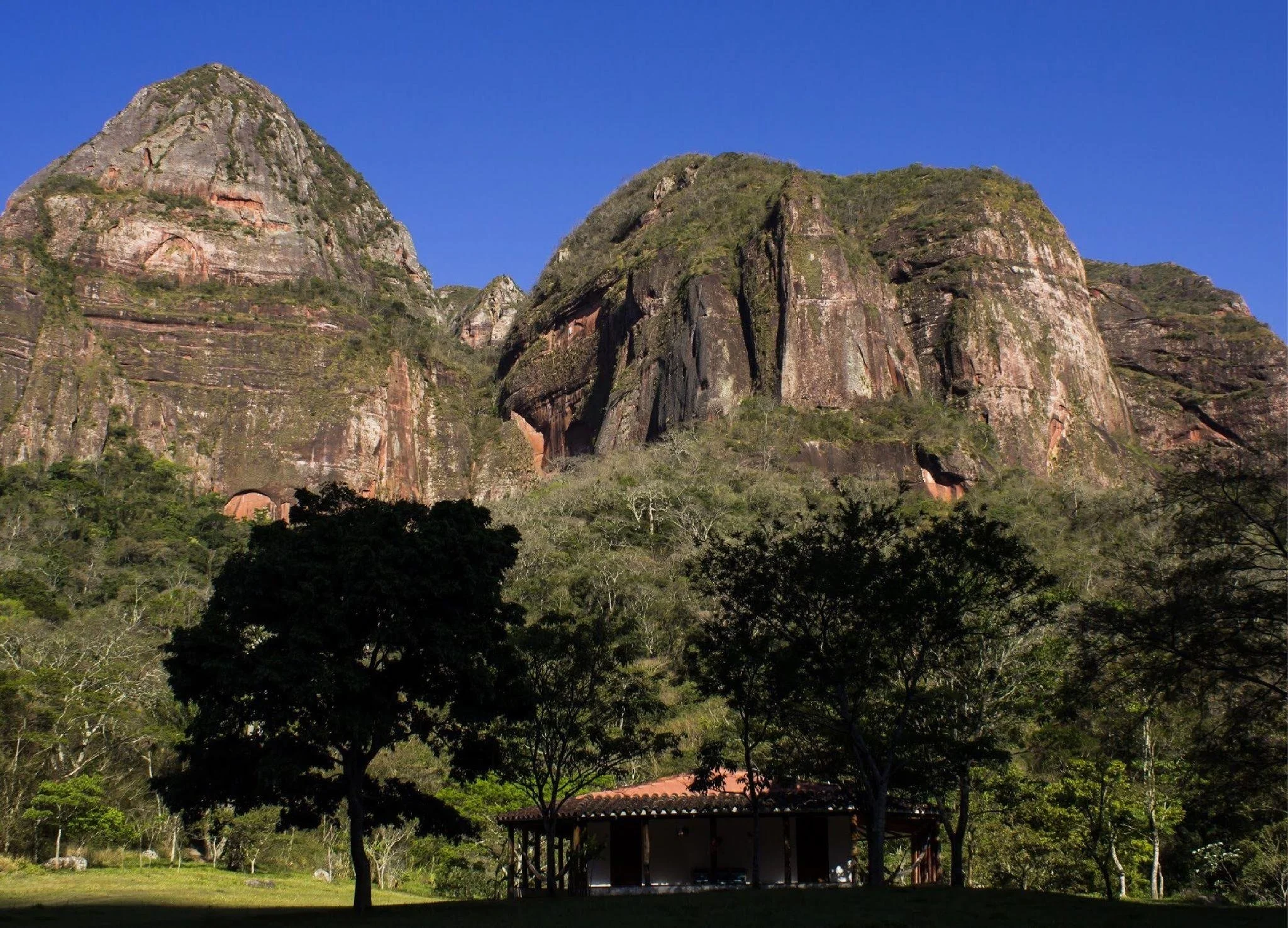 Berg mit Felsen und bewaldeter Basis, kleines Gebäude mit Terrasse im Vordergrund, blaue Himmel.