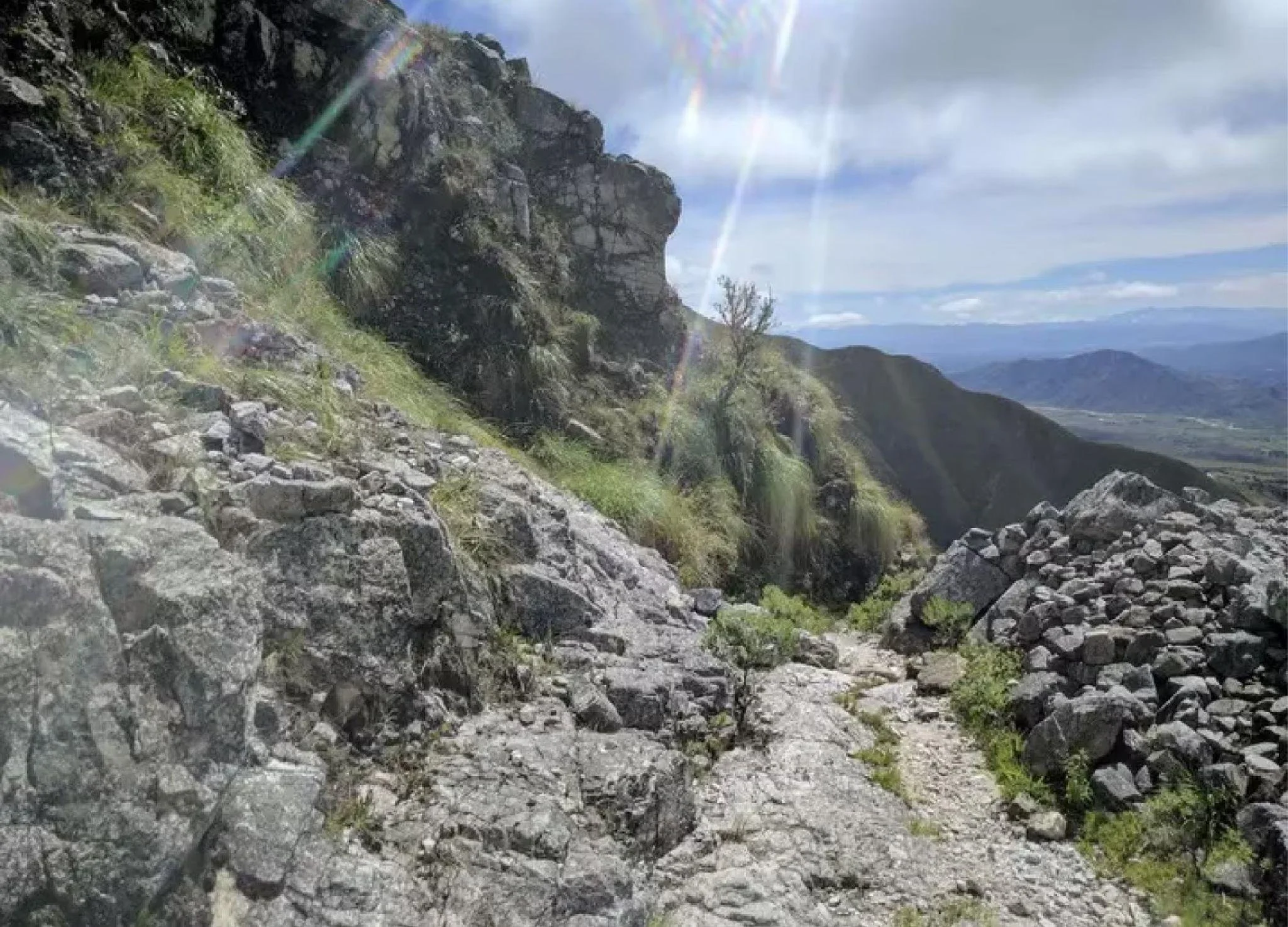 Bergpfad mit Steinen, umgeben von grüner Vegetation, Berglandschaft im Hintergrund, Sonnenstrahlen, blauer Himmel mit Wolken.