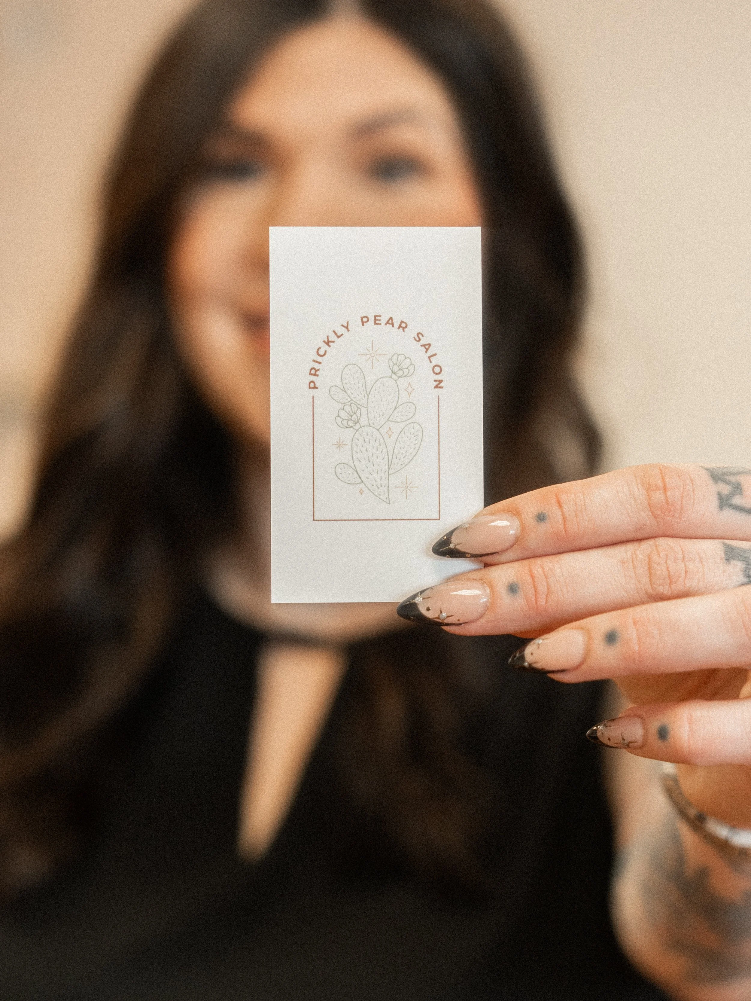 A woman holding a business card in front of her face, advertising Prickly Pear Salon with a cactus illustration.